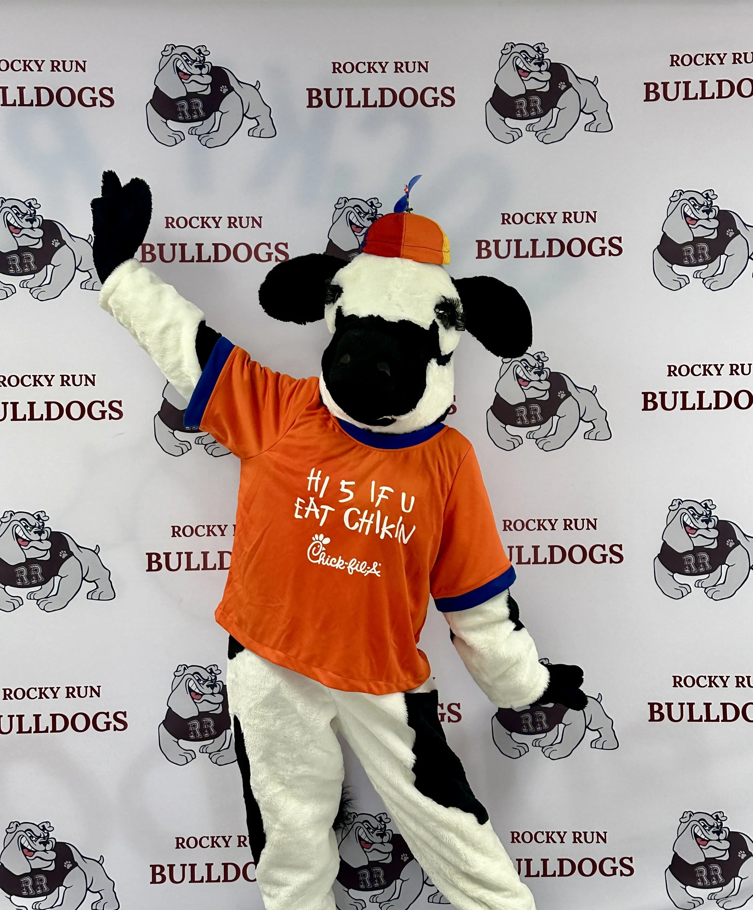 The Chick-fil-A Cow posing in front of a backdrop with the Rocky Run Bulldogs logo.