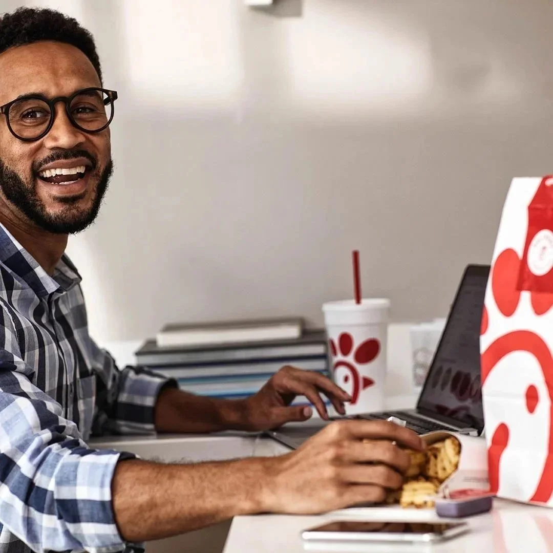Man eating Chick-fil-A sandwich at a desk with a laptop, drinks, and books.
