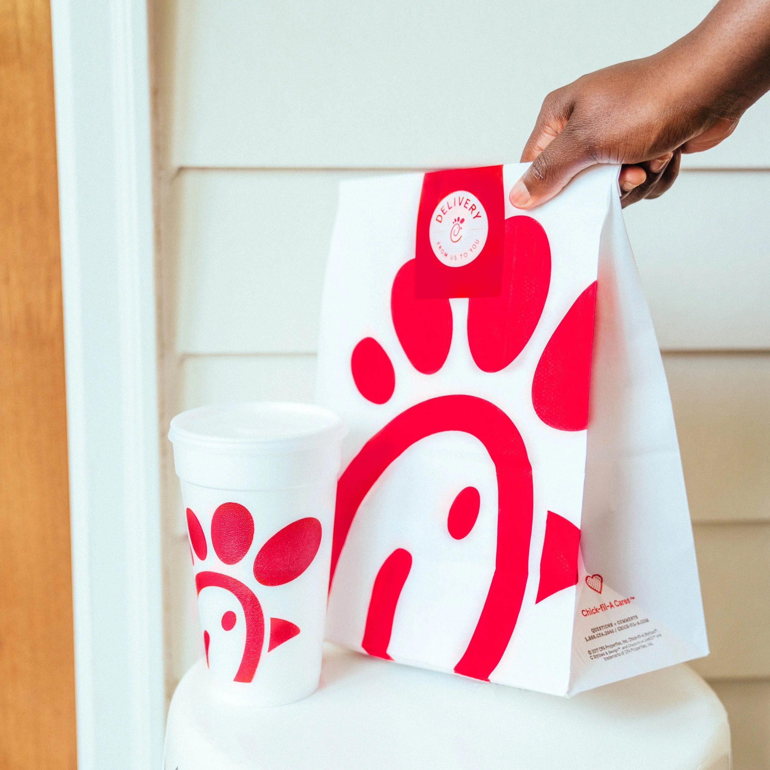 A hand holding a Chick-fil-A paper bag over a white cup on a table. The bag has the Chick-fil-A logo and the slogan 'Delivered right to you'.