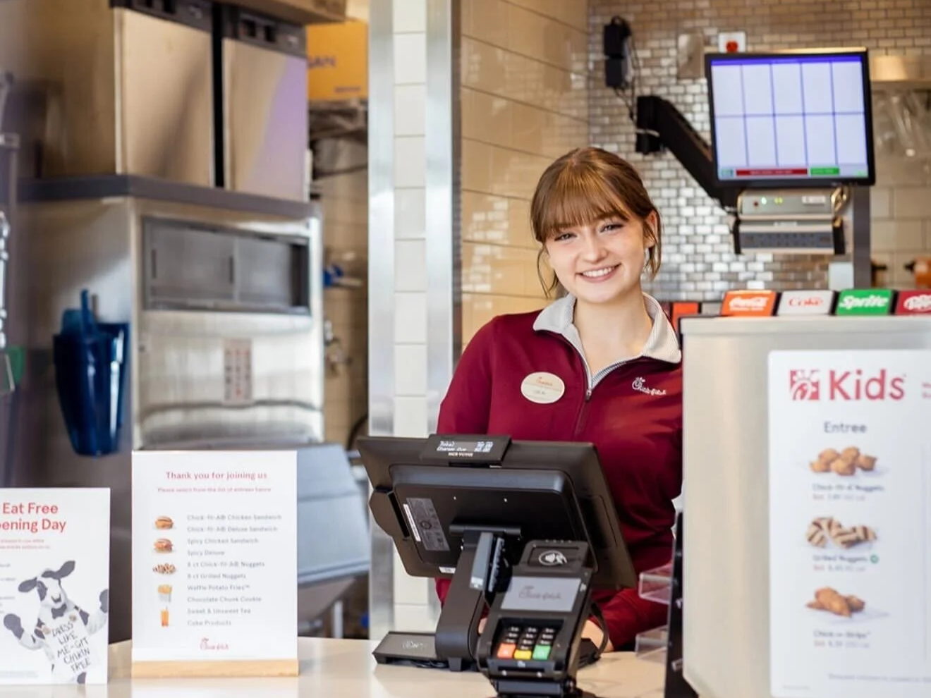 A smiling Chick-fil-A employee standing behind the counter at a restaurant, with a touchscreen register and menu displays visible.