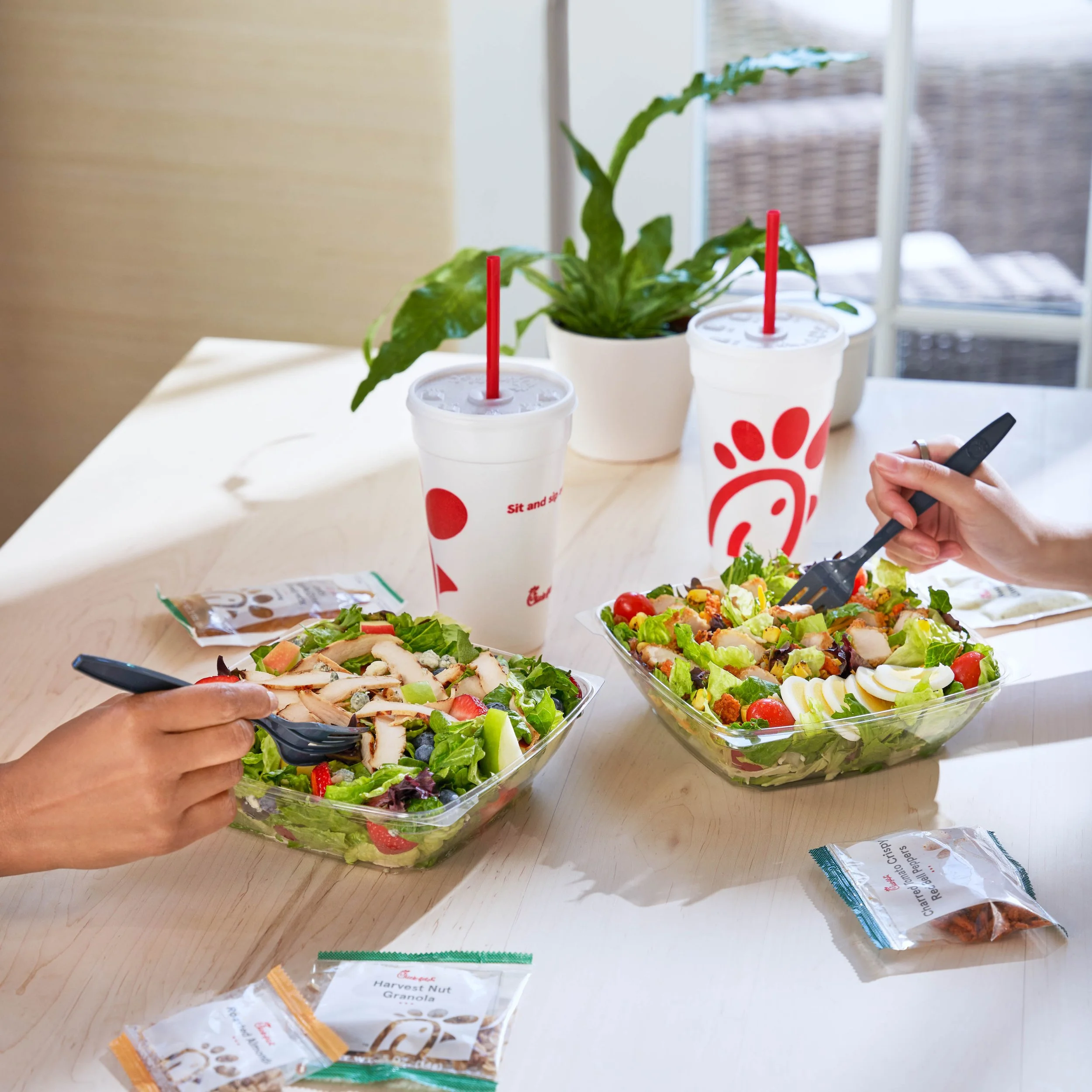 Three Chick-fil-A cups with red straws on a table, a potted plant in the background, and salads with greens, tomatoes, and sliced eggs.