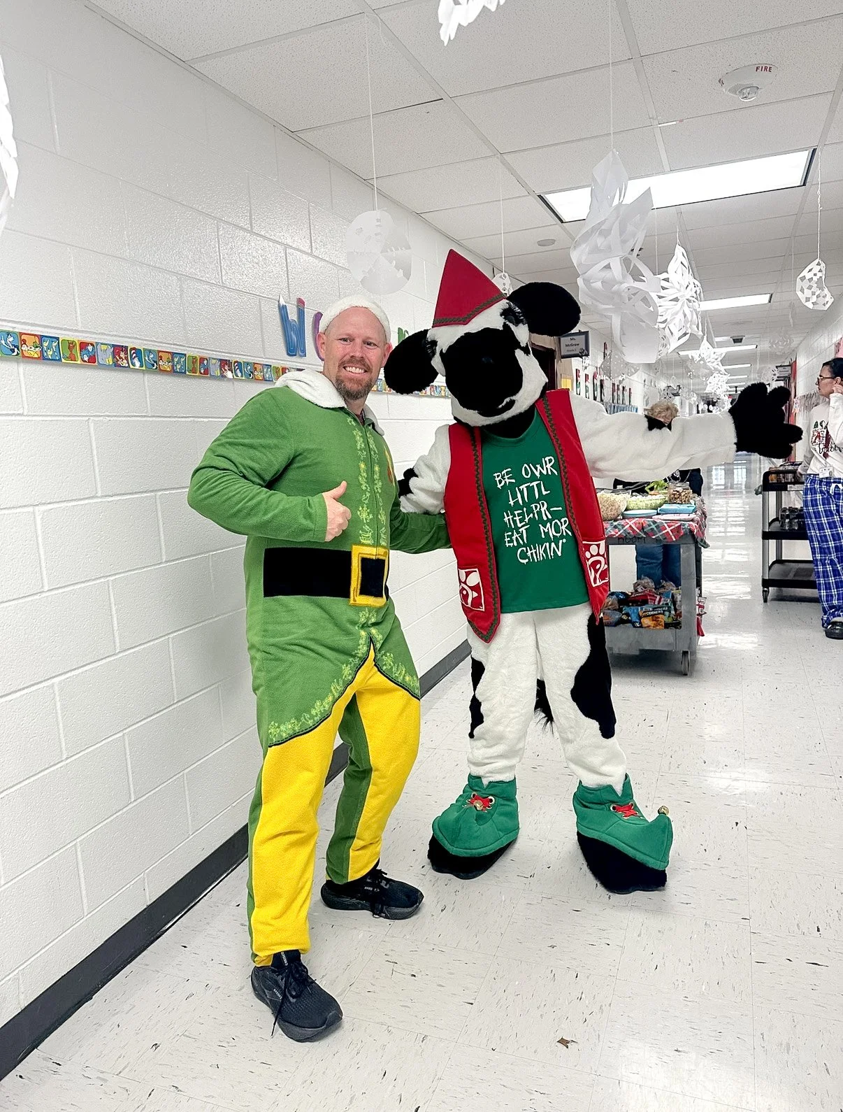 A man dressed as an elf standing next to a the Chick-fil-A Cow, both giving a thumbs-up in a decorated hallway with holiday ornaments.