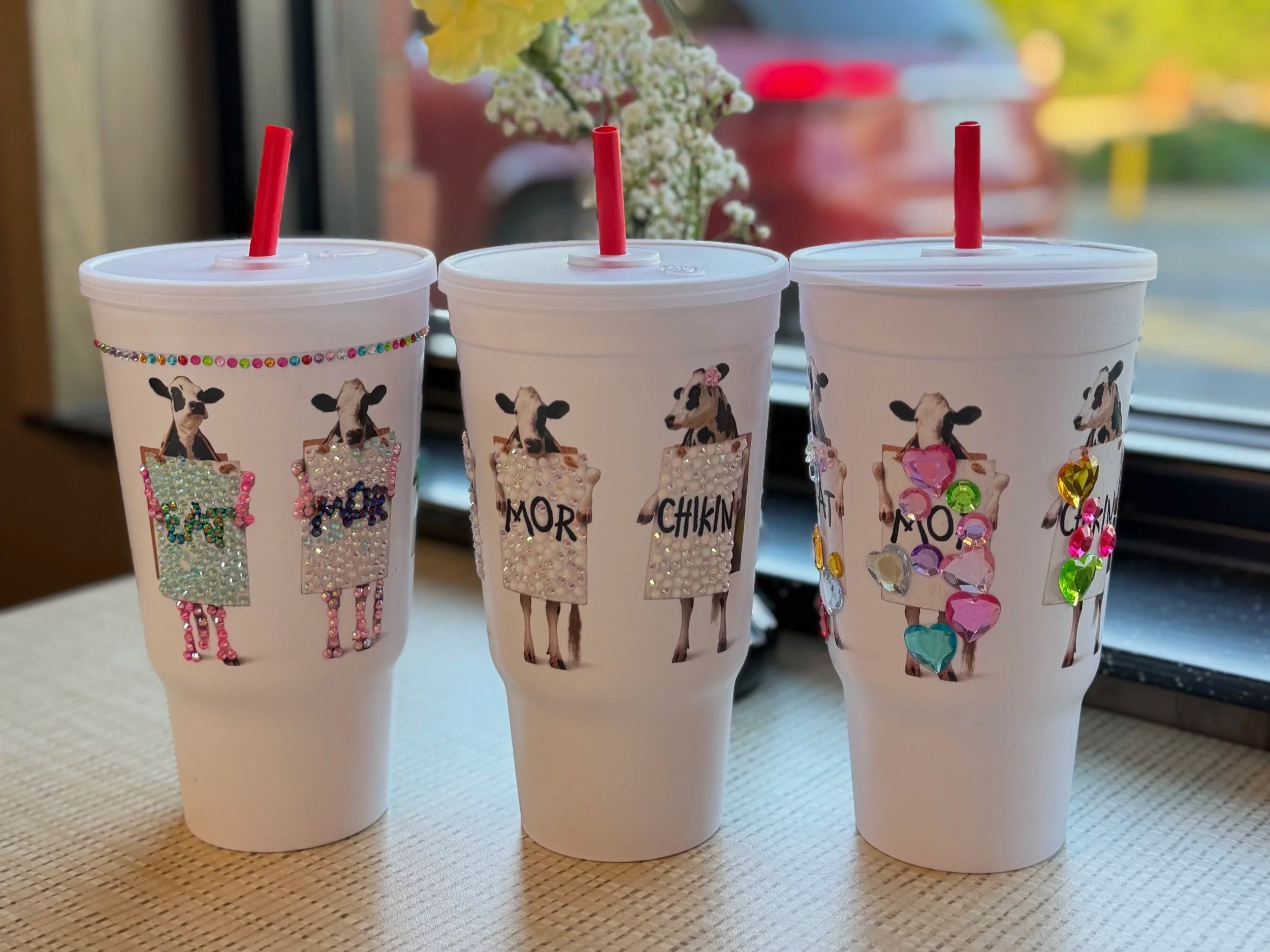 Three white disposable cups with animal designs and decorated with colorful rhinestones sitting on a table. Each cup has a red straw and labels that read 'MOM,' 'MORE,' and 'CHIKIN,' with a background of a window and a vase of flowers.