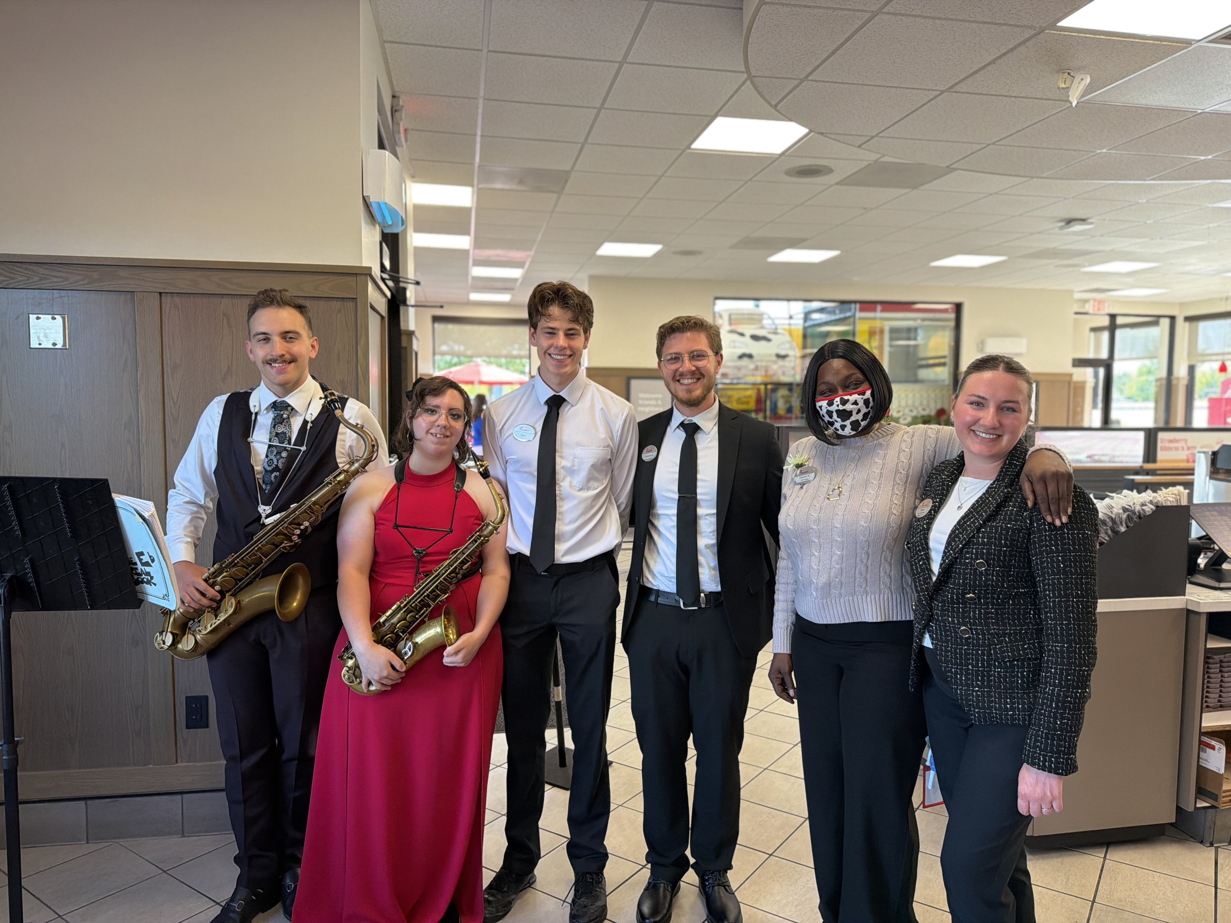 Group of six diverse people standing side by side inside a building, smiling at the camera. Two individuals are holding saxophones, and others are dressed casually or in business casual attire. The background shows a retail or service area with display shelves and windows.