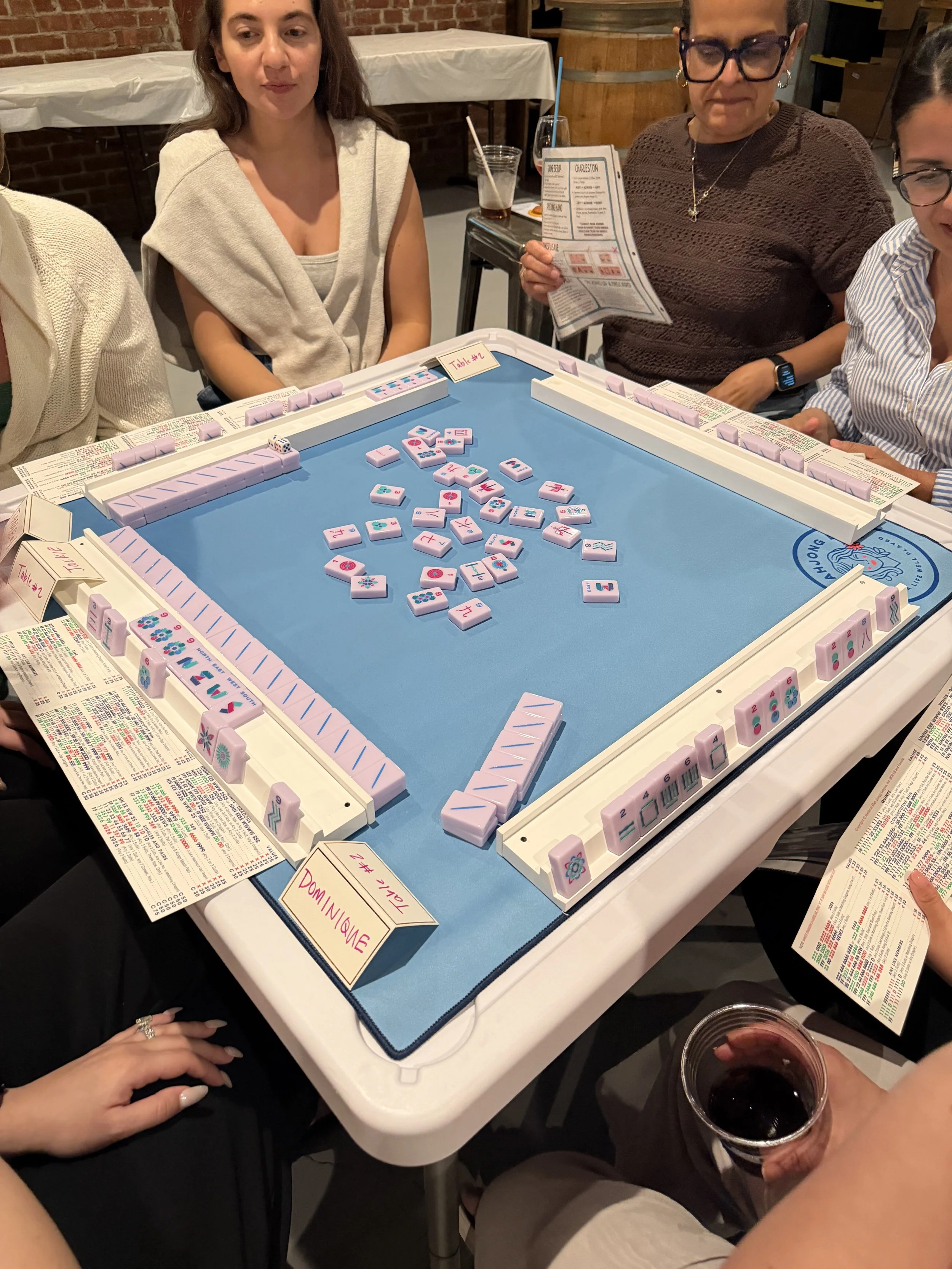Four women playing Mahjong at a table in a restaurant or bar, with drinks in hand, and a man's hand holding a glass of red wine visible in the foreground.