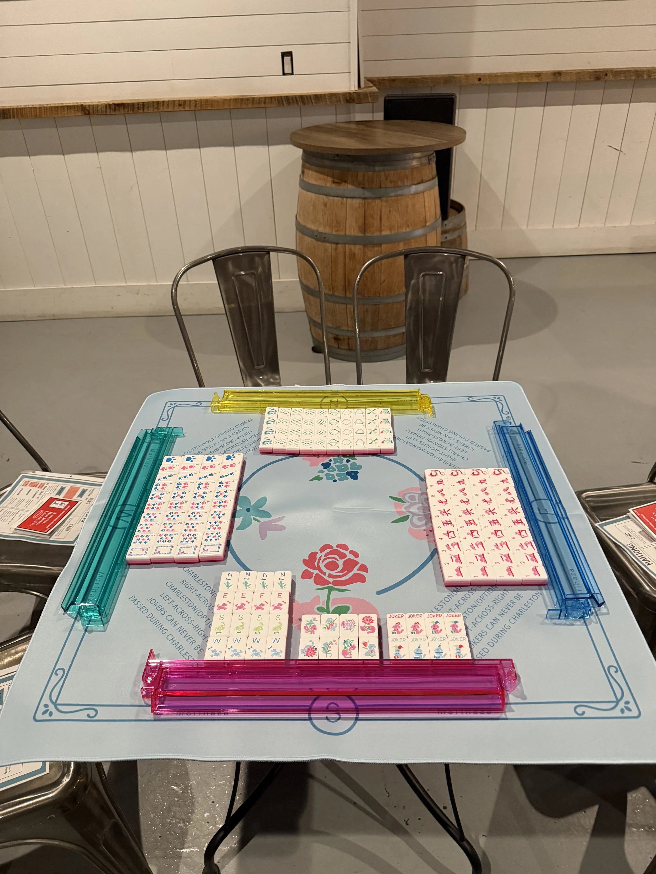 Table with Mahjong tiles, colorful plastic racks, and a floral design on the surface, set for a game of Mahjong. In the background, there is a wooden barrel-shaped table, metal chairs, and a white paneled wall.