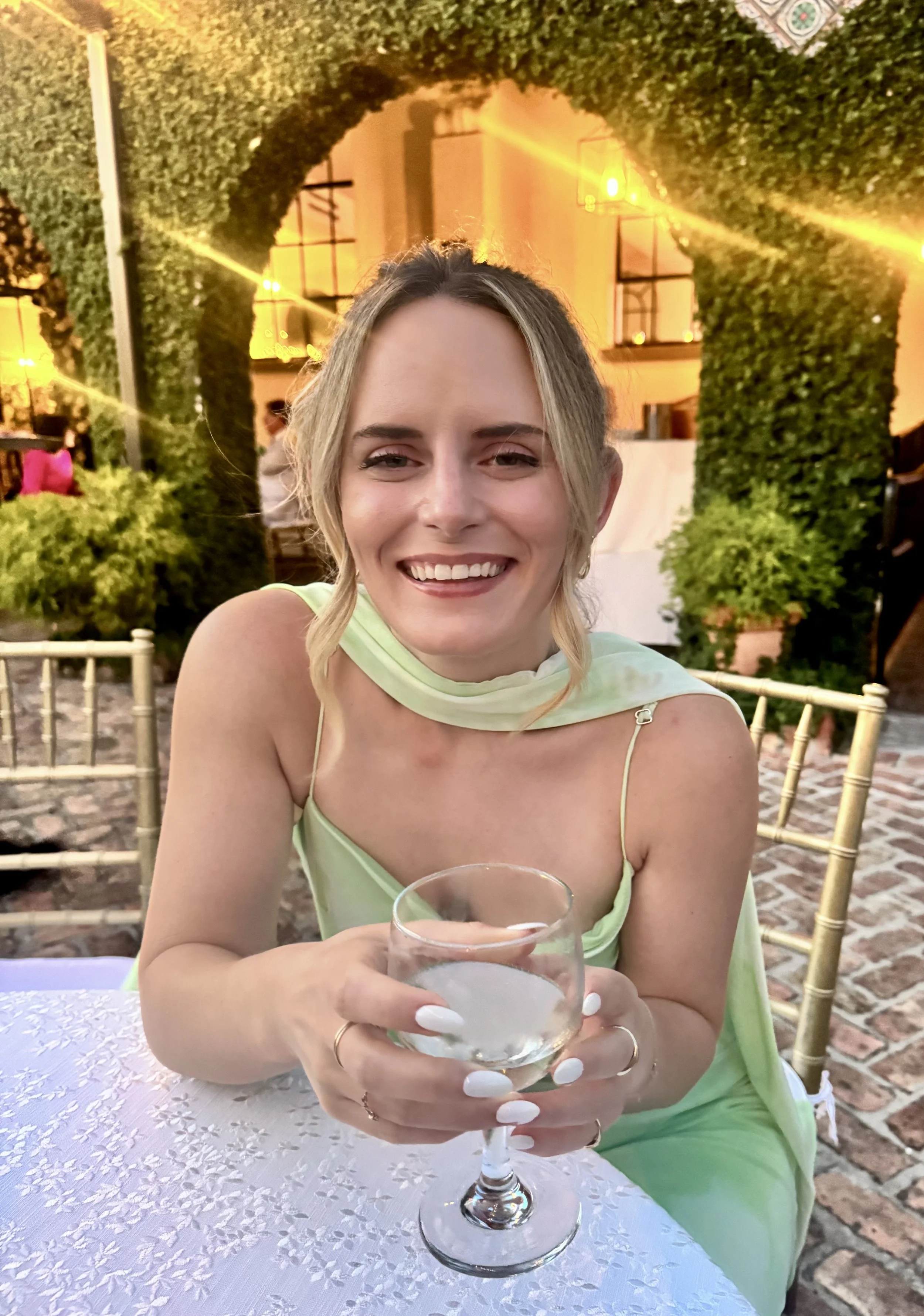 A woman in a light green dress sitting at a table, holding a glass of wine, smiling at the camera, with a decorative green hedge arch and warm outdoor lighting in the background.