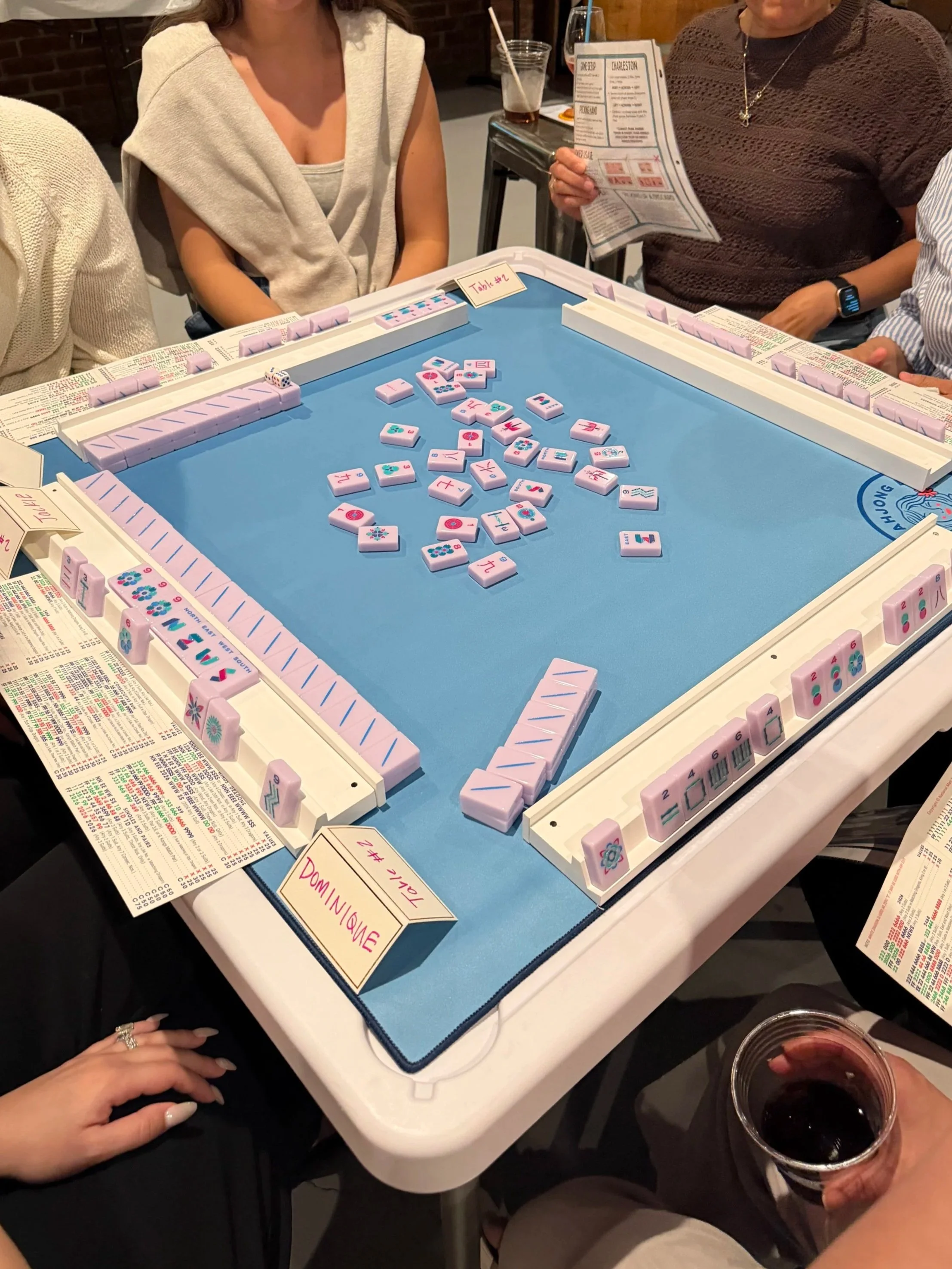 People playing dominoes at a table with drinks, including glasses of dark beverages, at a social gathering. The dominoes are pink with decorative symbols, and there are handwritten signs on the table indicating game details.