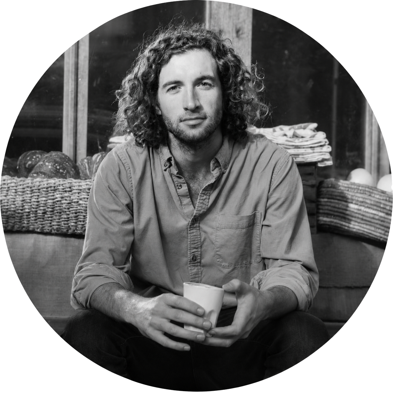 A young man with curly hair and a beard sitting indoors, holding a mug, with a basket of bread and stacks of paper or napkins behind him.