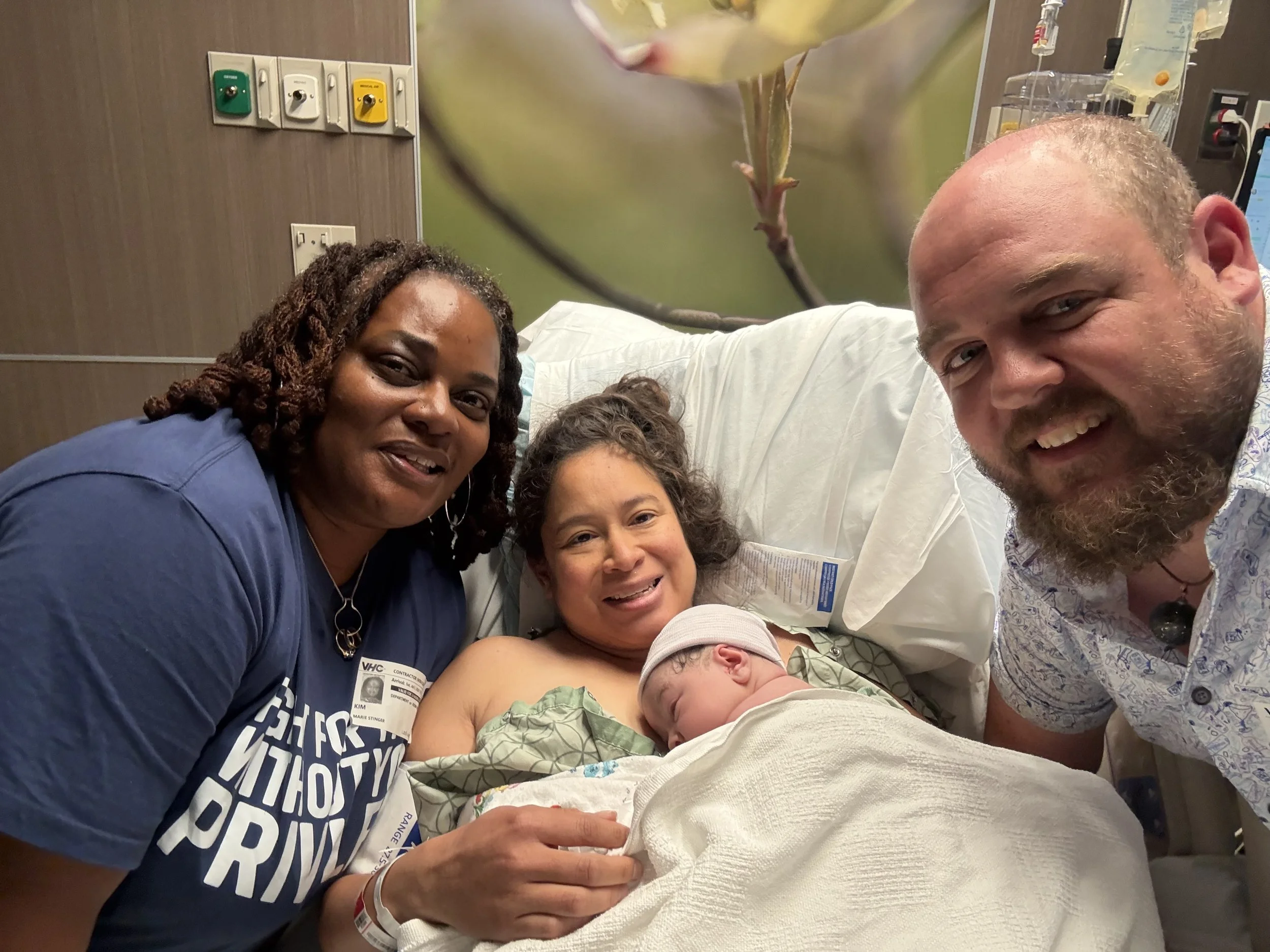 A group of five people, including children and adults, in a hospital room, smiling and taking a selfie with a newborn baby.