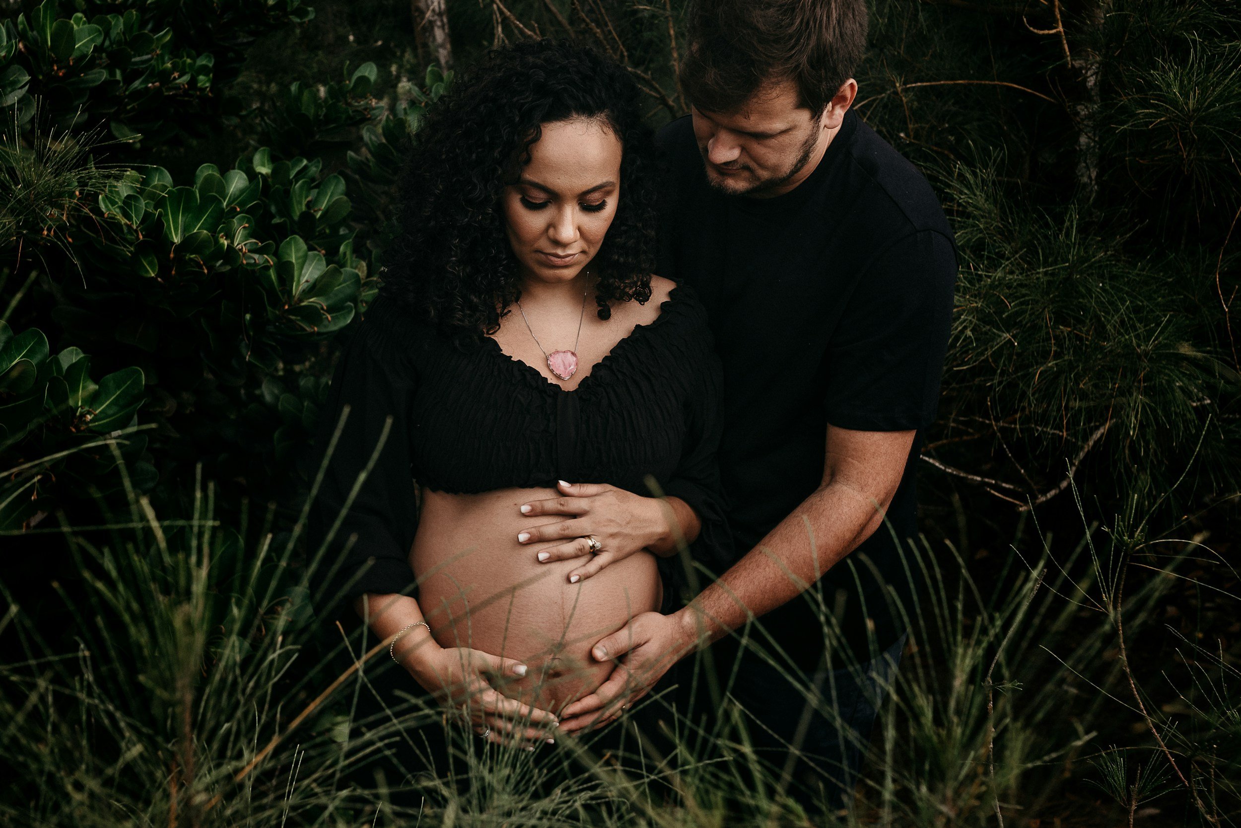 A couple in a lush green outdoor setting, with the woman holding her pregnant belly and the man embracing her from behind, both with serious expressions.