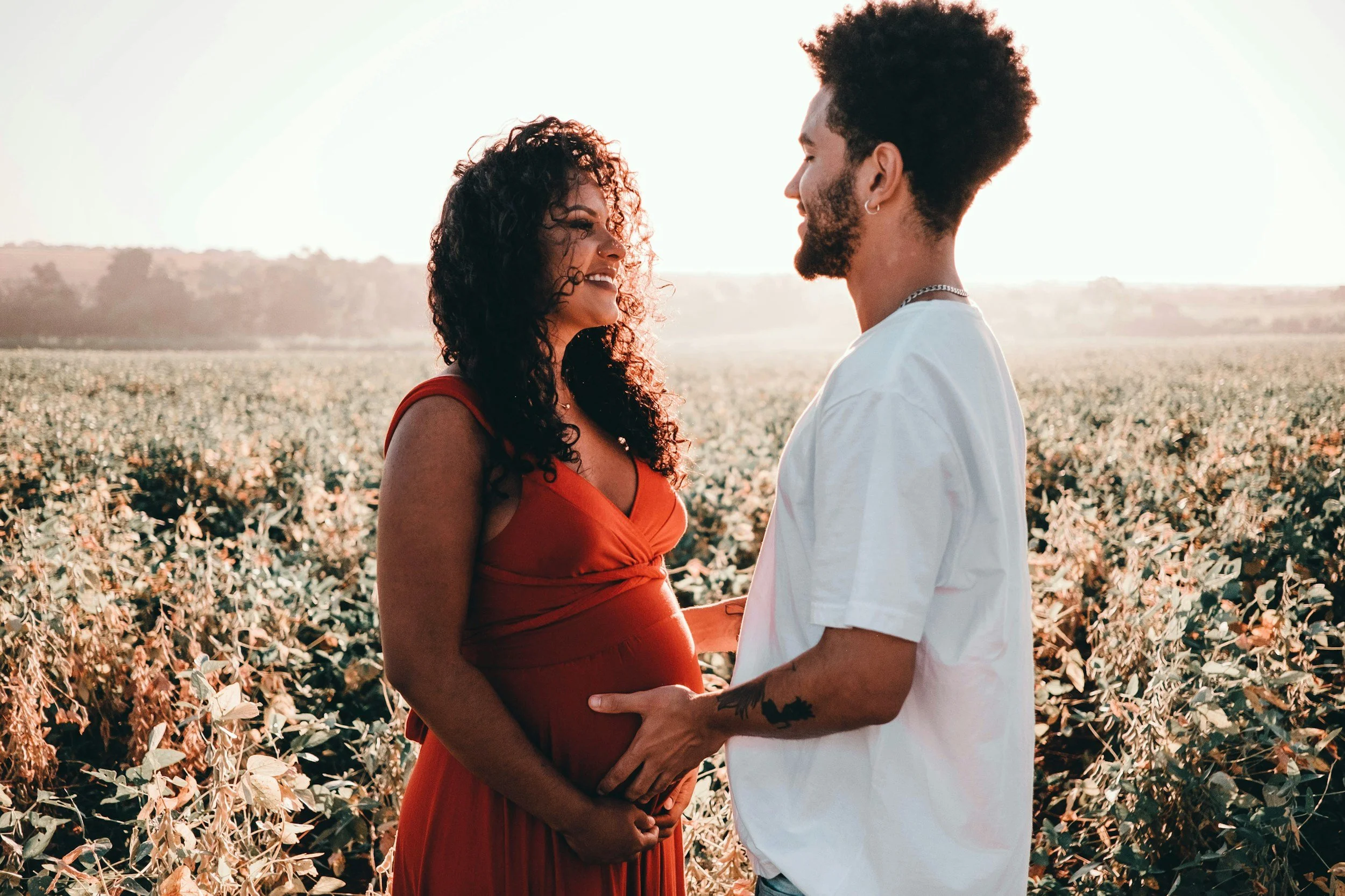 A couple standing in a field, facing each other and smiling, with the sunset in the background.