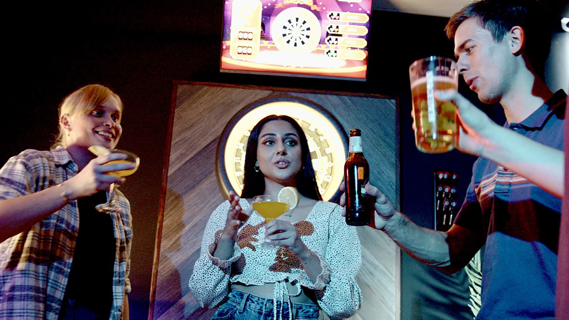 Three young adults having drinks and socializing at a bar or nightclub. There is a woman in the center holding a cocktail and a bottle, a woman on the left holding a glass, and a man on the right holding a beer, with a dartboard and a screen in the background.