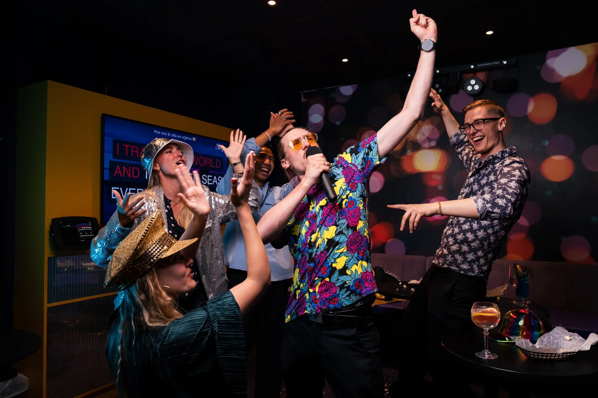 A group of diverse people singing and dancing at a karaoke night, with a woman in colorful floral shirt holding a microphone and others with joyful expressions.