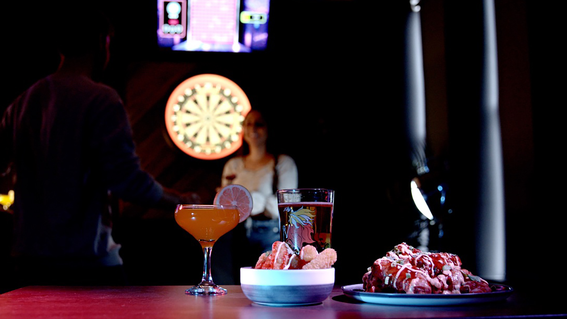 Bar scene with two cocktails on the table, one garnished with a citrus slice, and plates of desserts. In the background, a man and a woman are near the dartboard and blue-lit screens.