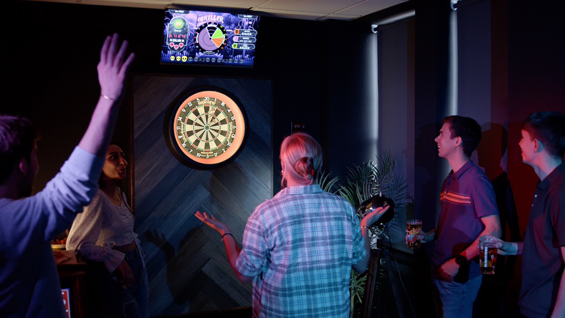 Group of people gathered in an indoor space playing darts, with a dartboard on the wall, some holding drinks, and a small screen above the dartboard displaying game information.