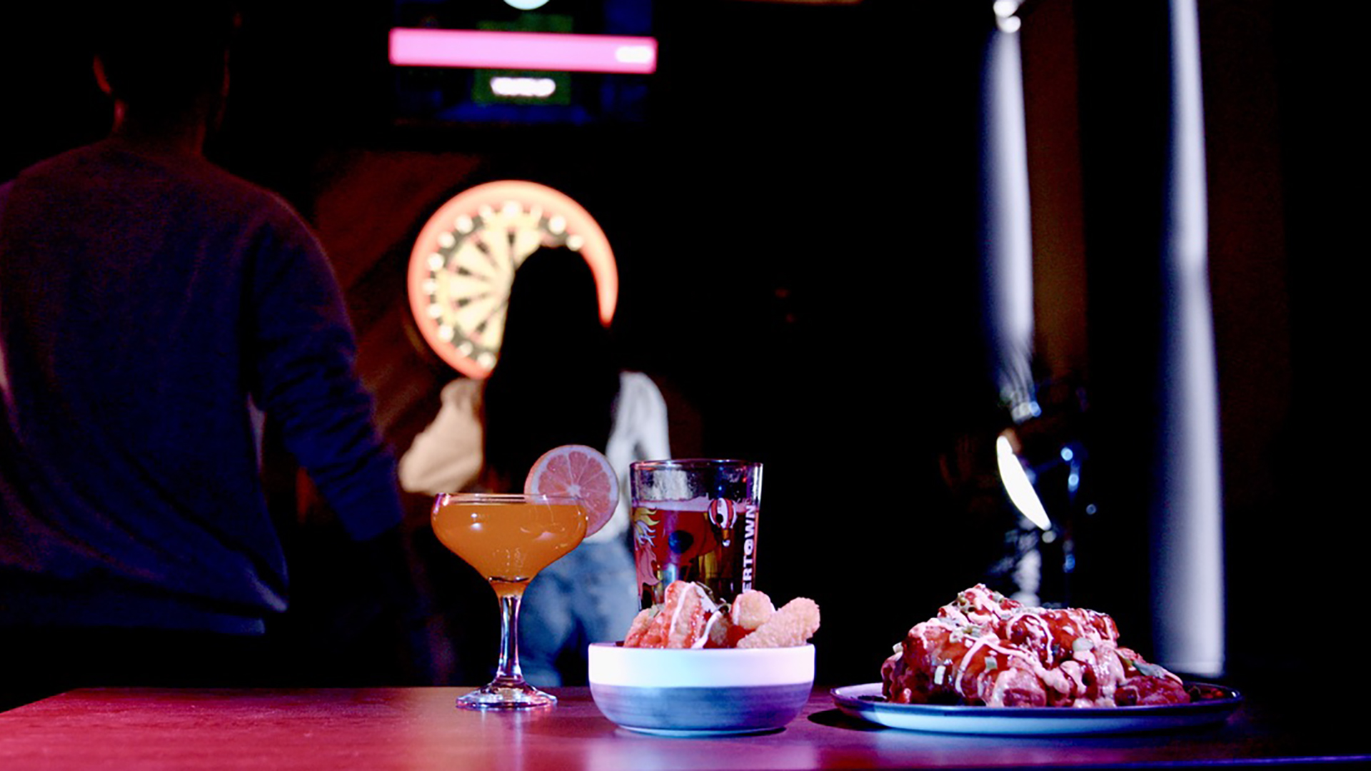 Cocktail glasses and plates of dessert on a bar counter with people in the background, illuminated by low lighting and neon signs.