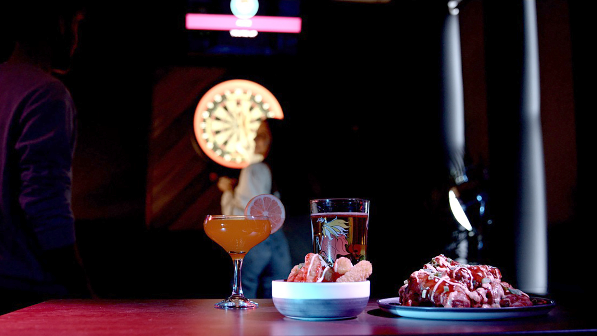 Two cocktails, one with a lemon slice and the other with a lemon twist, on a table in a dark bar. In the background, a woman standing near a dartboard, with a dart in her hand, and a man off to the left side of the image.
