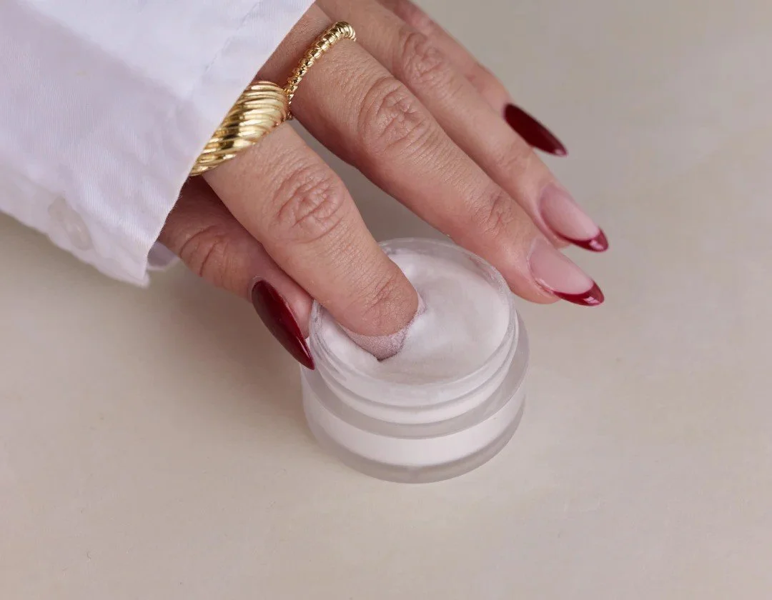 Hand with red nails dipping into a small jar of white cream.