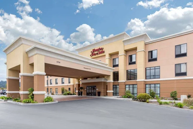 View of a Hampton Inn hotel with a covered entrance, beige and peach-colored exterior, surrounded by landscaping, under a partly cloudy sky.