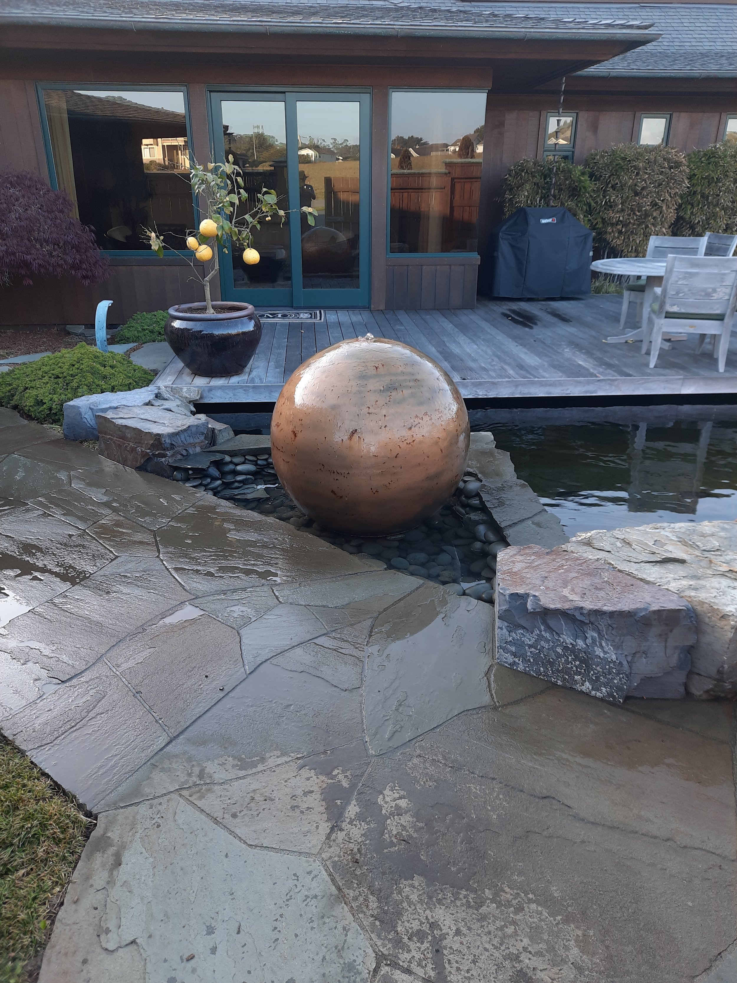 An outdoor backyard area featuring a wooden deck with patio furniture, a covered grill, a potted lemon tree, a decorative water feature with a large spherical fountain, and stone paving leading to a grassy patch.