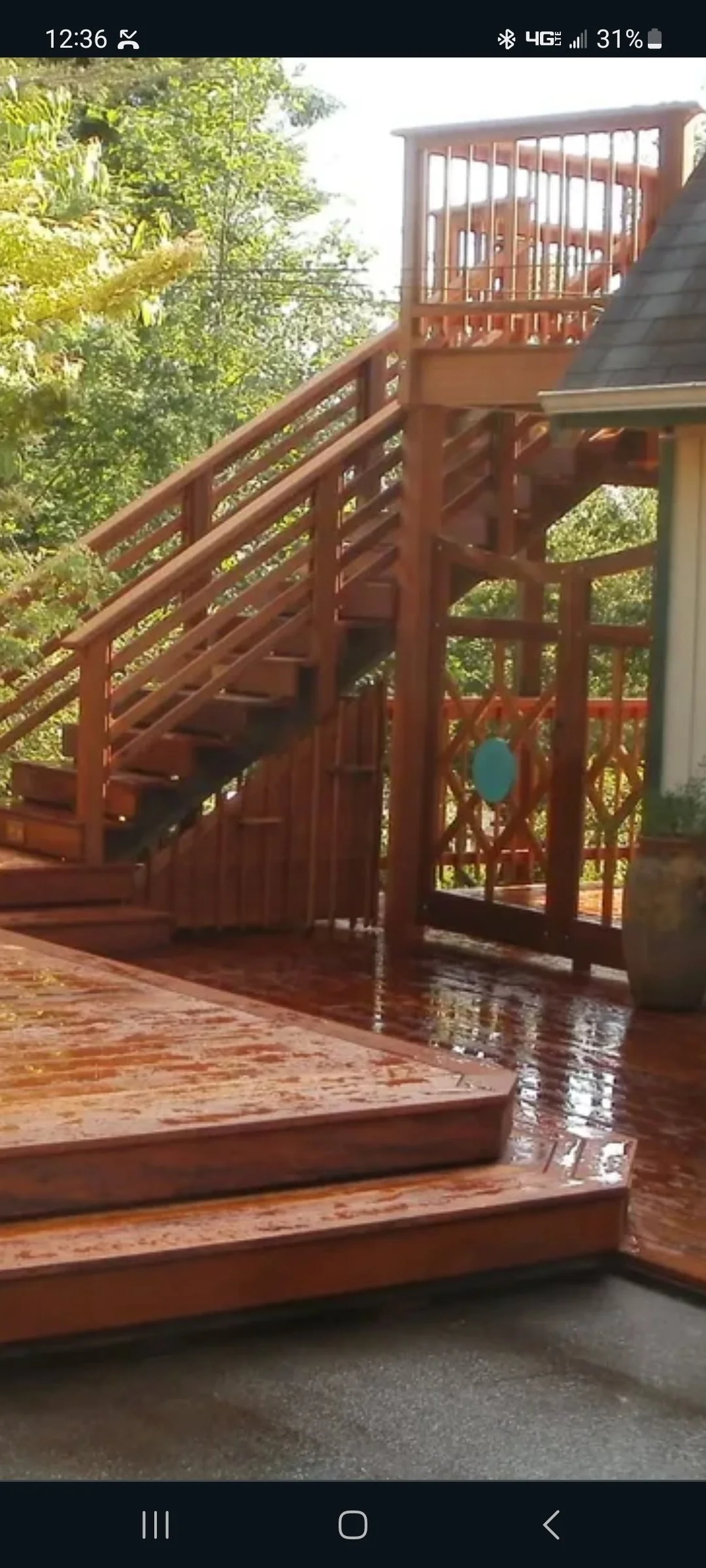 Wooden outdoor staircase with a landing leading to a second level, surrounded by lush green trees, on a wet wooden deck.