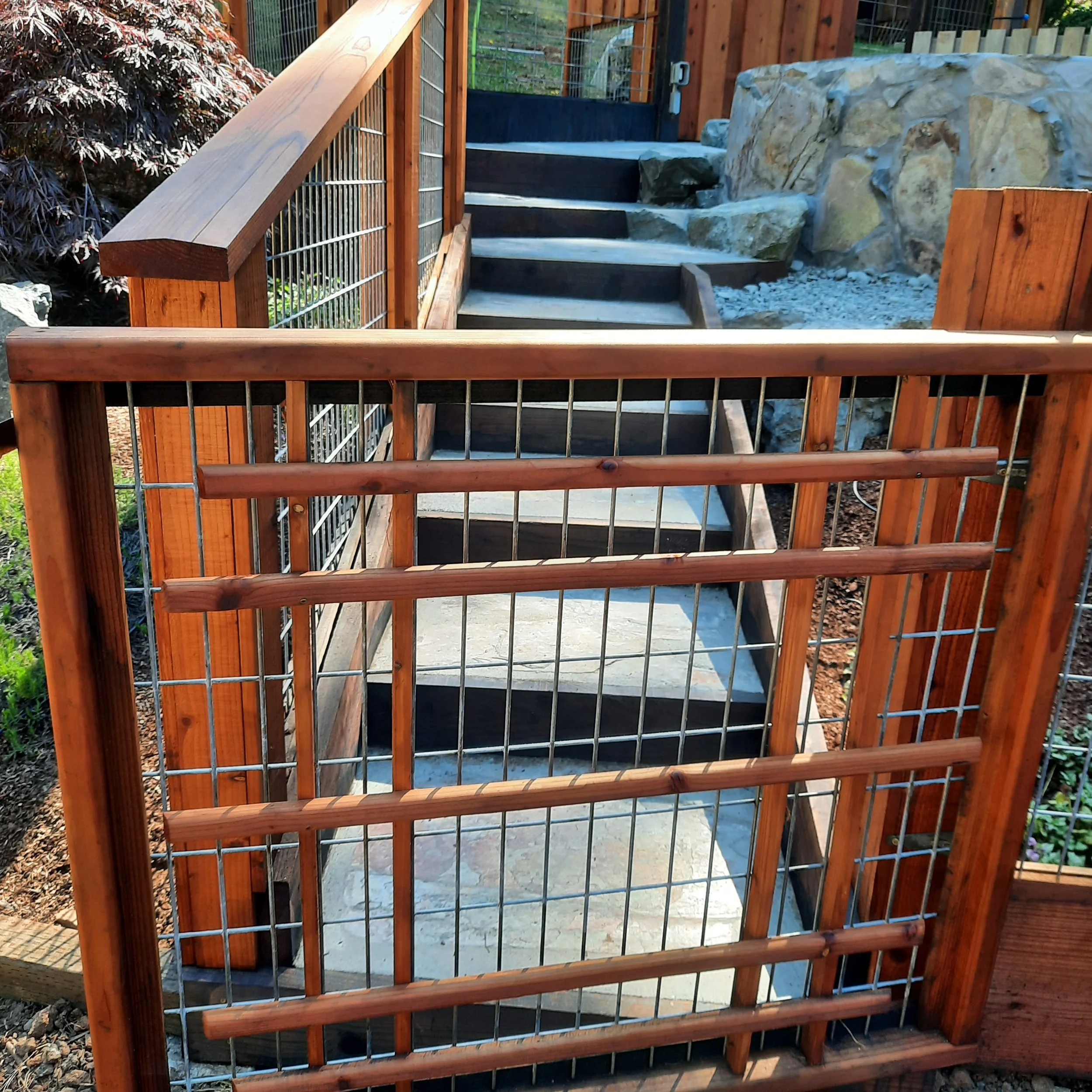 Wooden staircase with metal railings leading up to a black door, next to a large rock hot tub and surrounded by landscaping including a tree with dark red leaves.