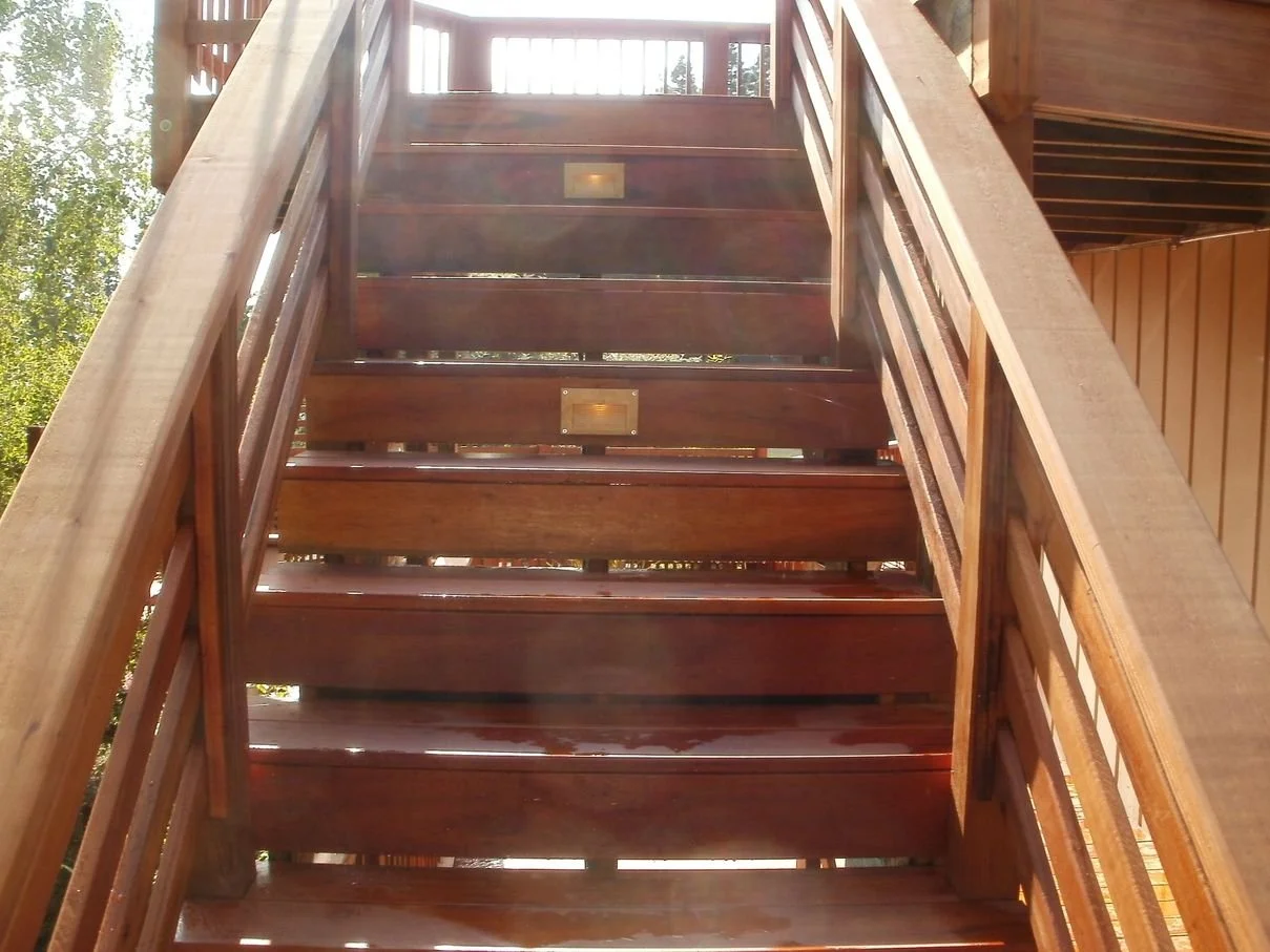 exotic wood outdoor staircase with handrails, stairs and inset lights leading up, sunlight streaming through, and trees visible in the background.