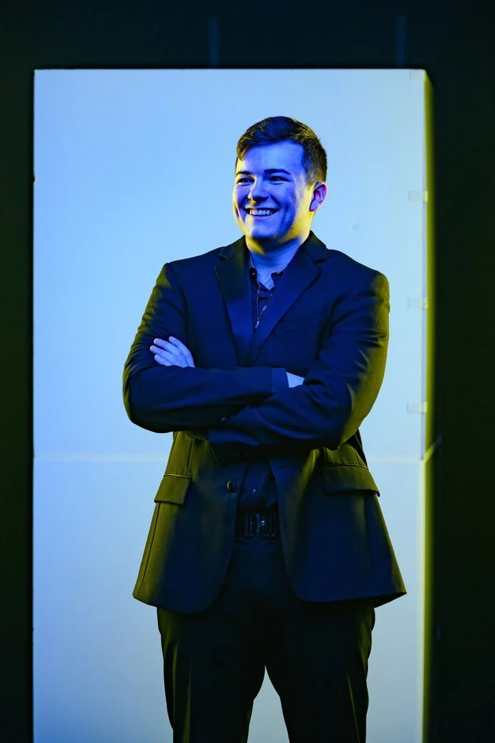 A young man with dark hair, dressed in a dark suit, smiling and crossing his arms, standing in front of a plain, light blue screen in a dark room.