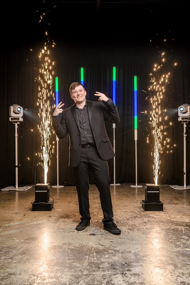 A young man in a black suit making peace signs with both hands, standing on a stage decorated with colorful light bars and surrounded by sparklers erupting on either side, in a celebratory setting.