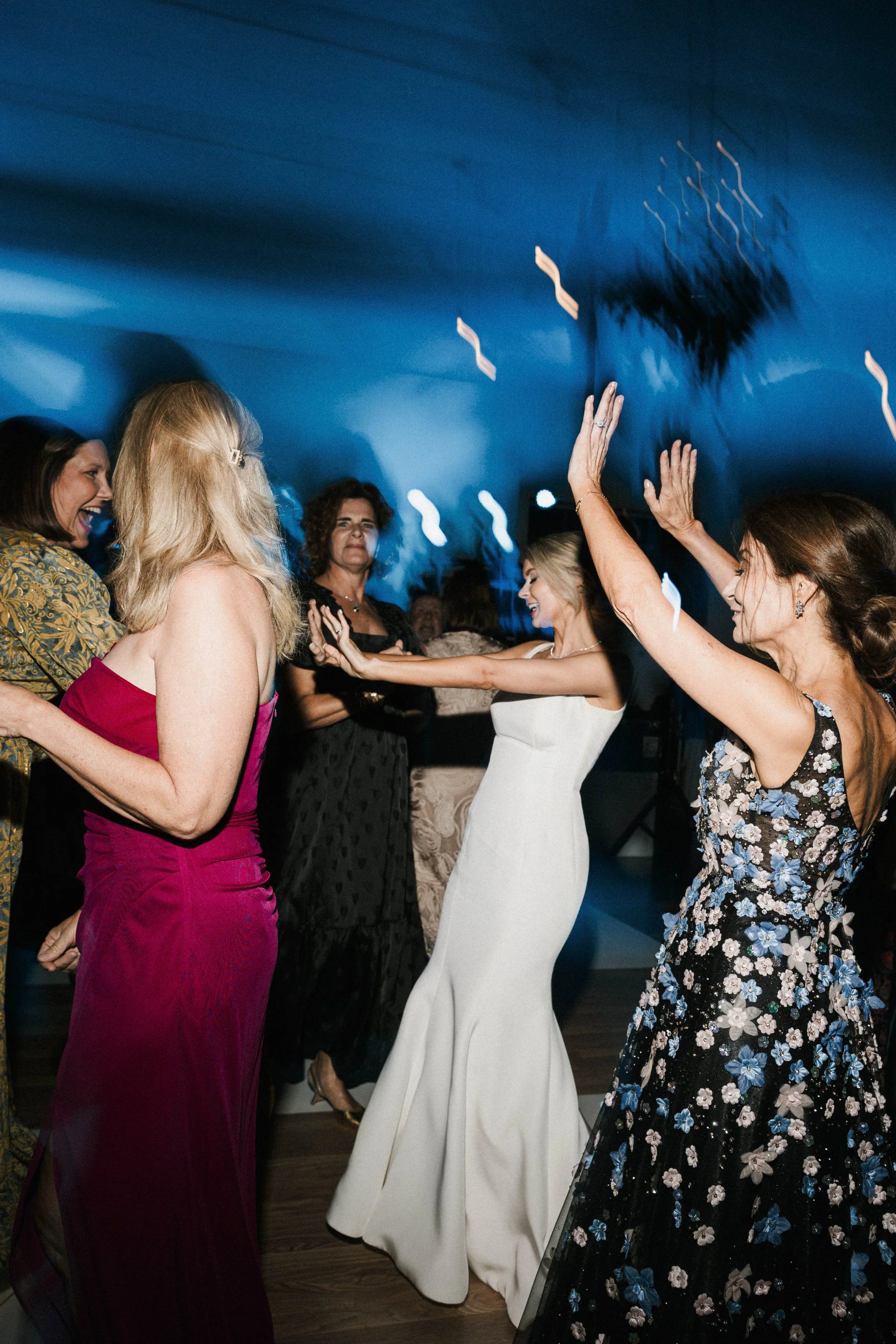 Group of women dancing at a party or wedding celebration, smiling and raising their hands, with a blue ceiling and dim lighting.