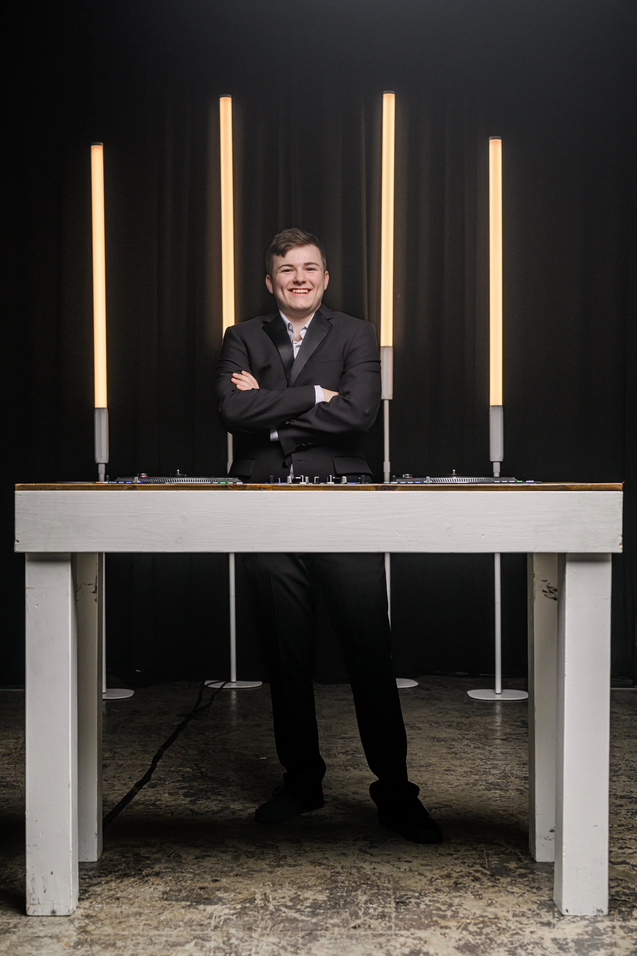 Smiling young man in a black suit with arms crossed, standing behind a DJ table with turntables, four vertical light tubes in the background, black curtain backdrop, and wooden flooring.