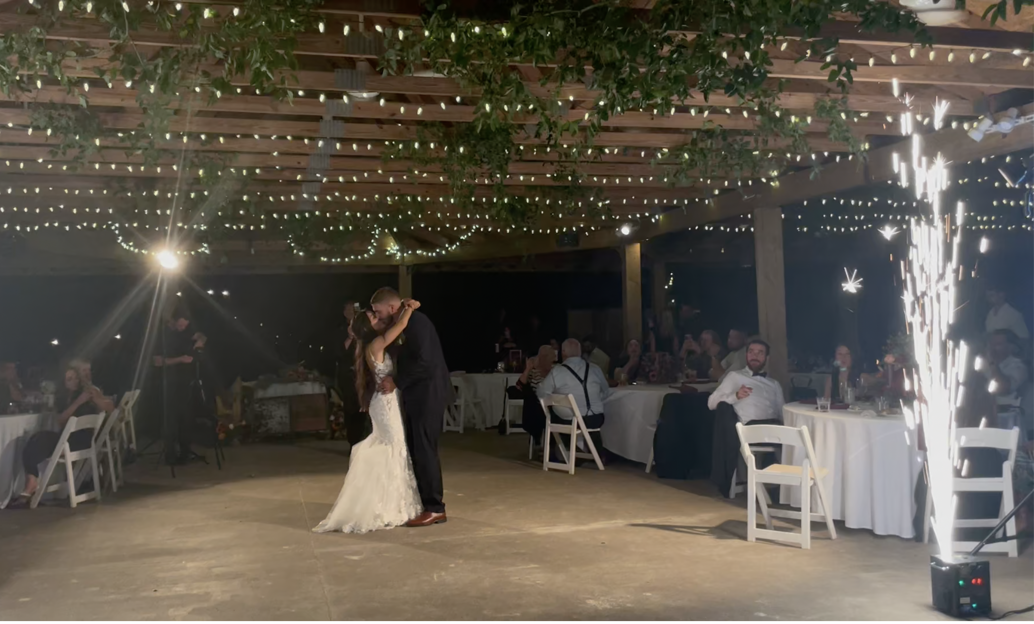 A newlywed couple shares a dance at their wedding reception during the evening, under string lights and surrounded by seated guests and a sparkler with firework display.