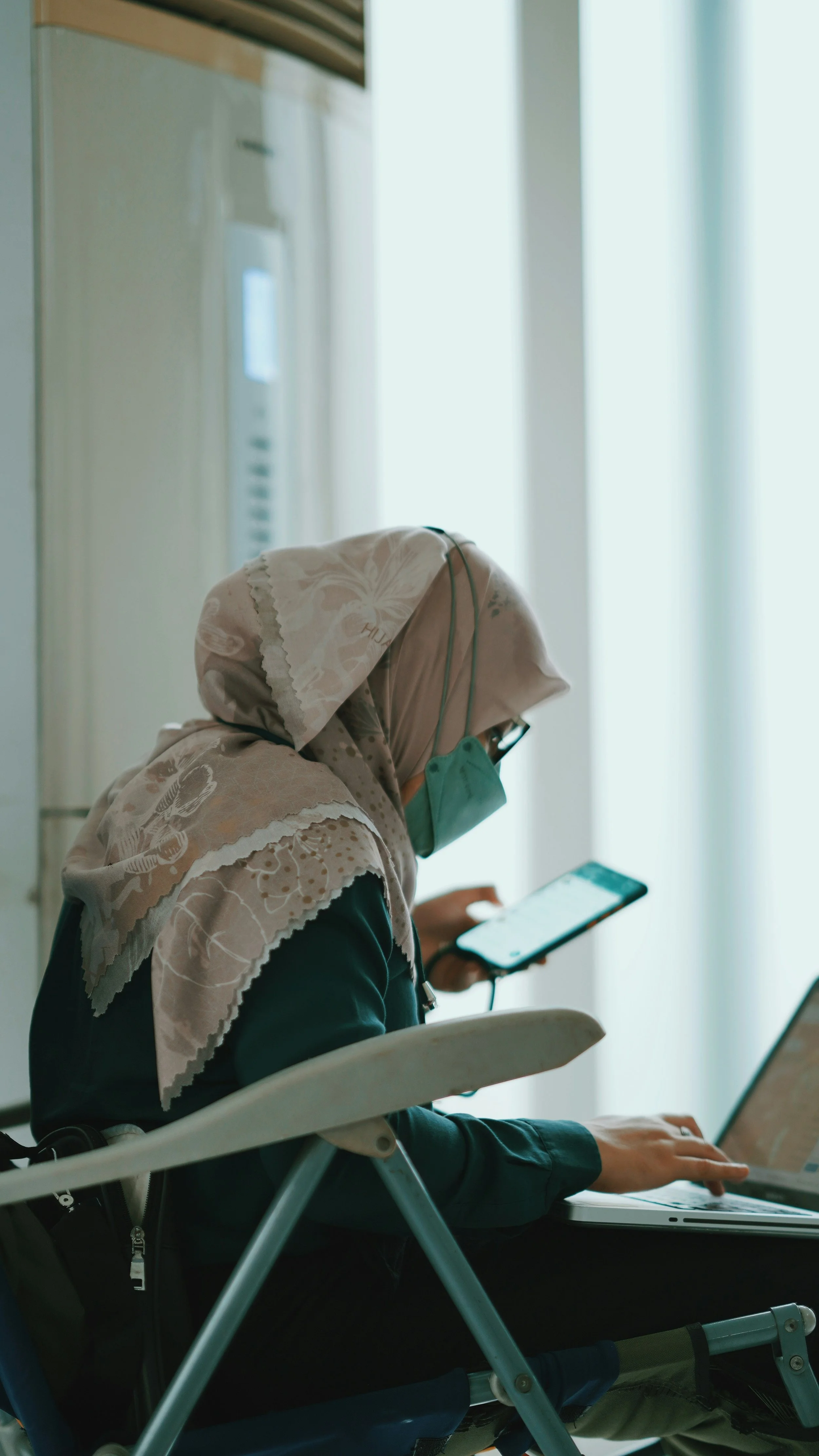 A woman wearing a headscarf and a face mask, sitting in a white chair, looking at her smartphone, with a laptop on her lap, in a bright room with large windows.