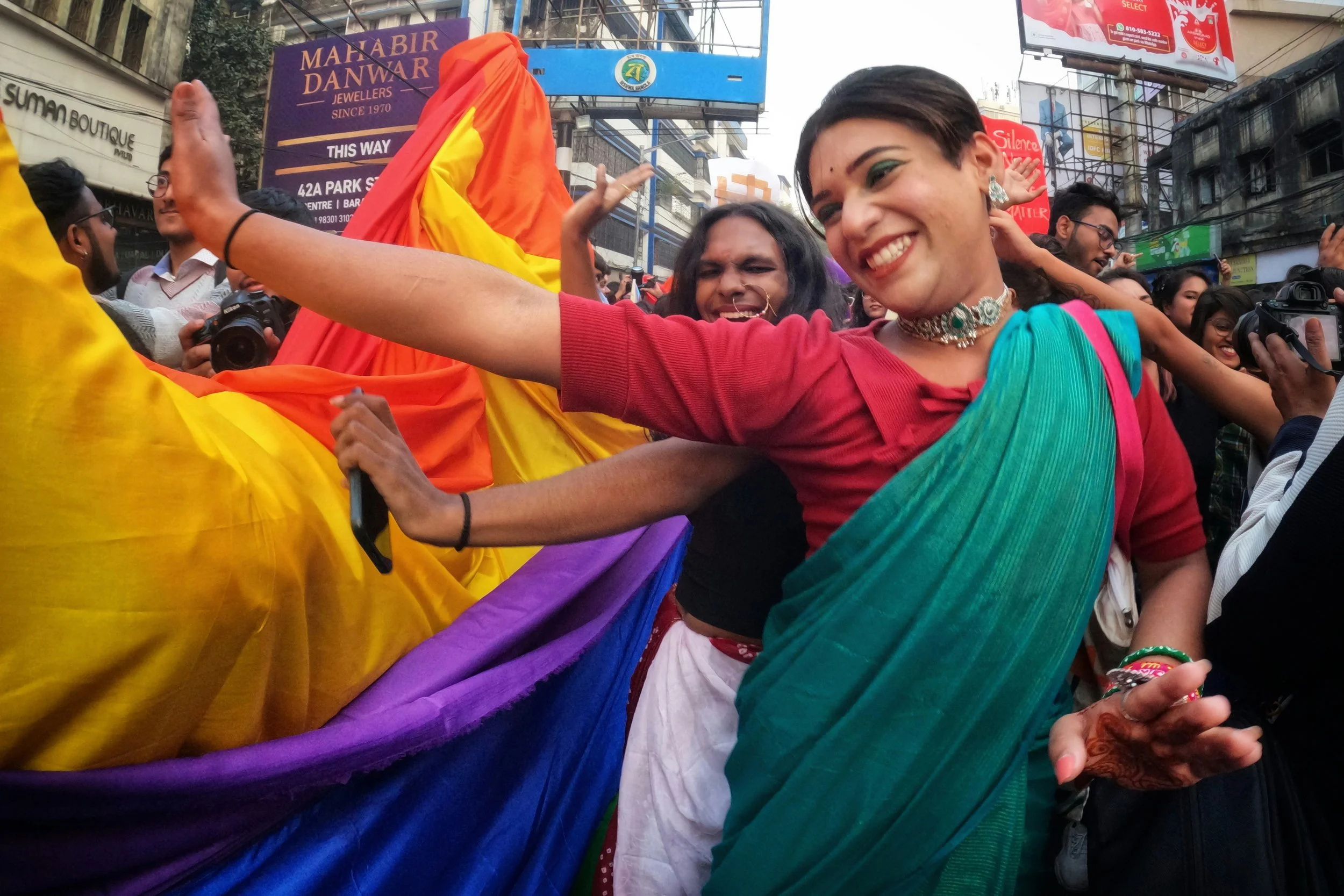 People celebrating on a street with rainbow fabric, smiling and taking photos, during a pride parade or event.