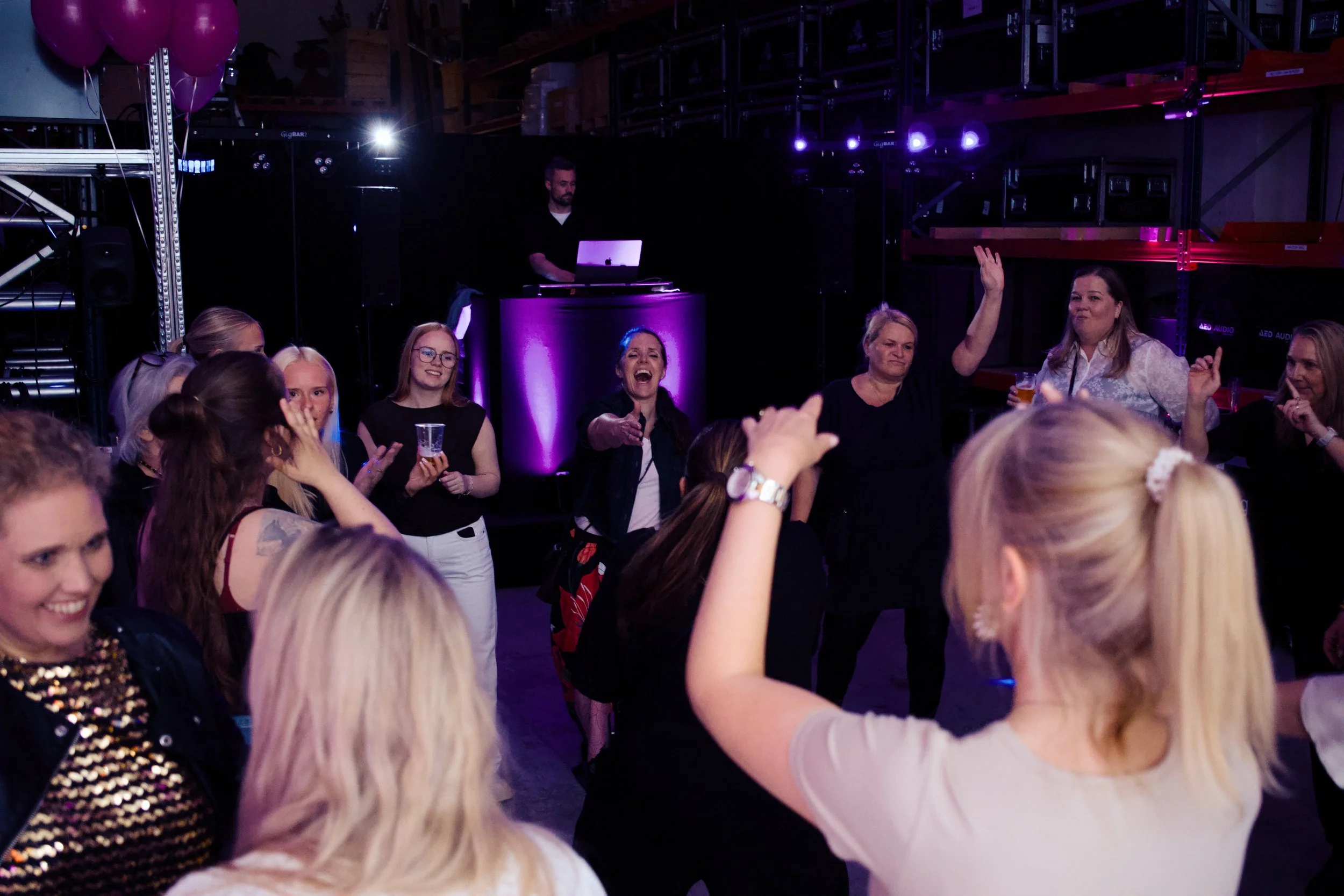 People dancing and celebrating at a party or concert with a DJ playing music in the background, purple lighting, and balloons.