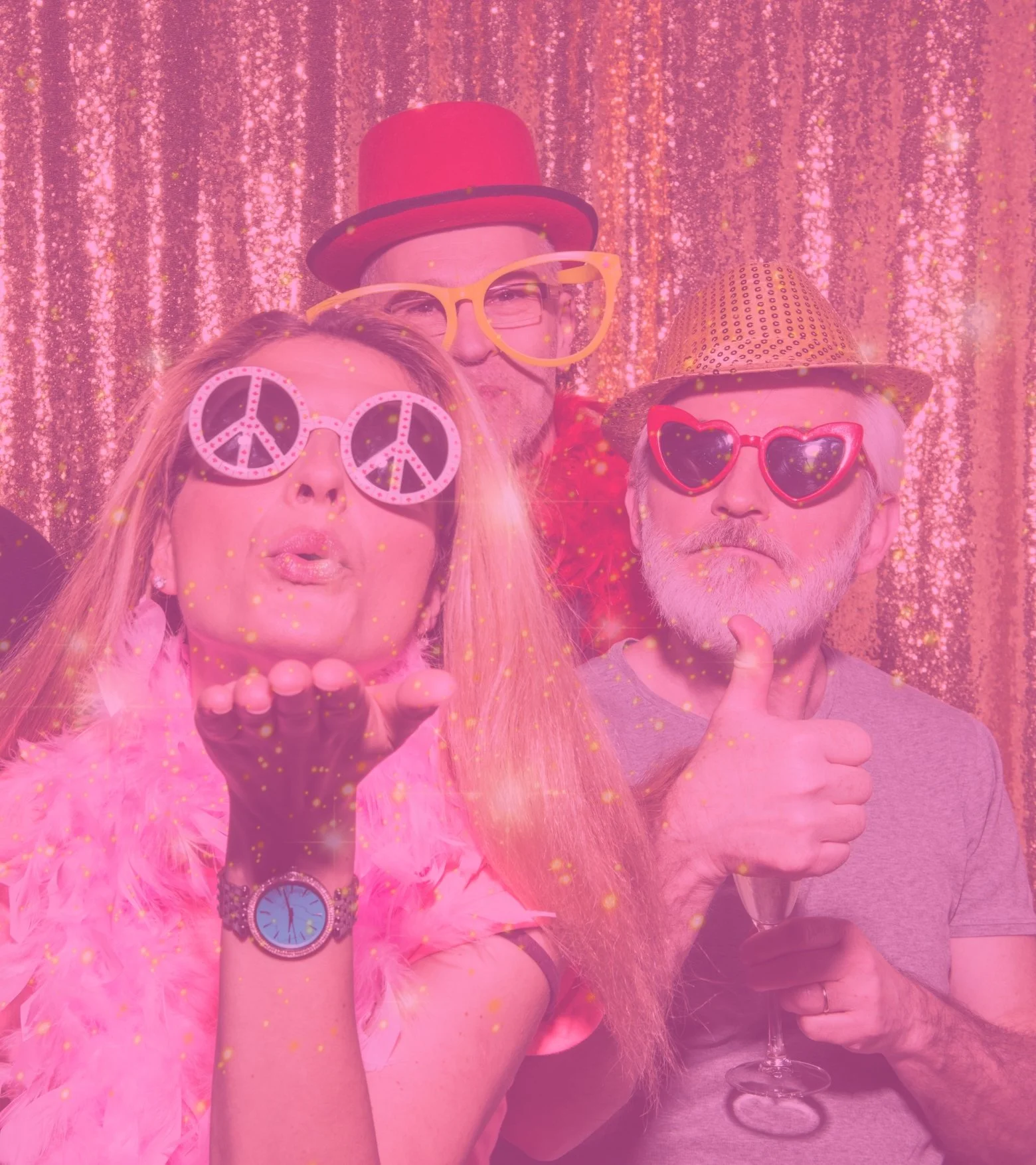 Three people dressed in colorful, fun accessories, standing in front of a glittery, pink background; one person wearing pink peace sign sunglasses, another with yellow glasses and a top hat, and the third with heart-shaped glasses and a straw hat, celebrating at a party.