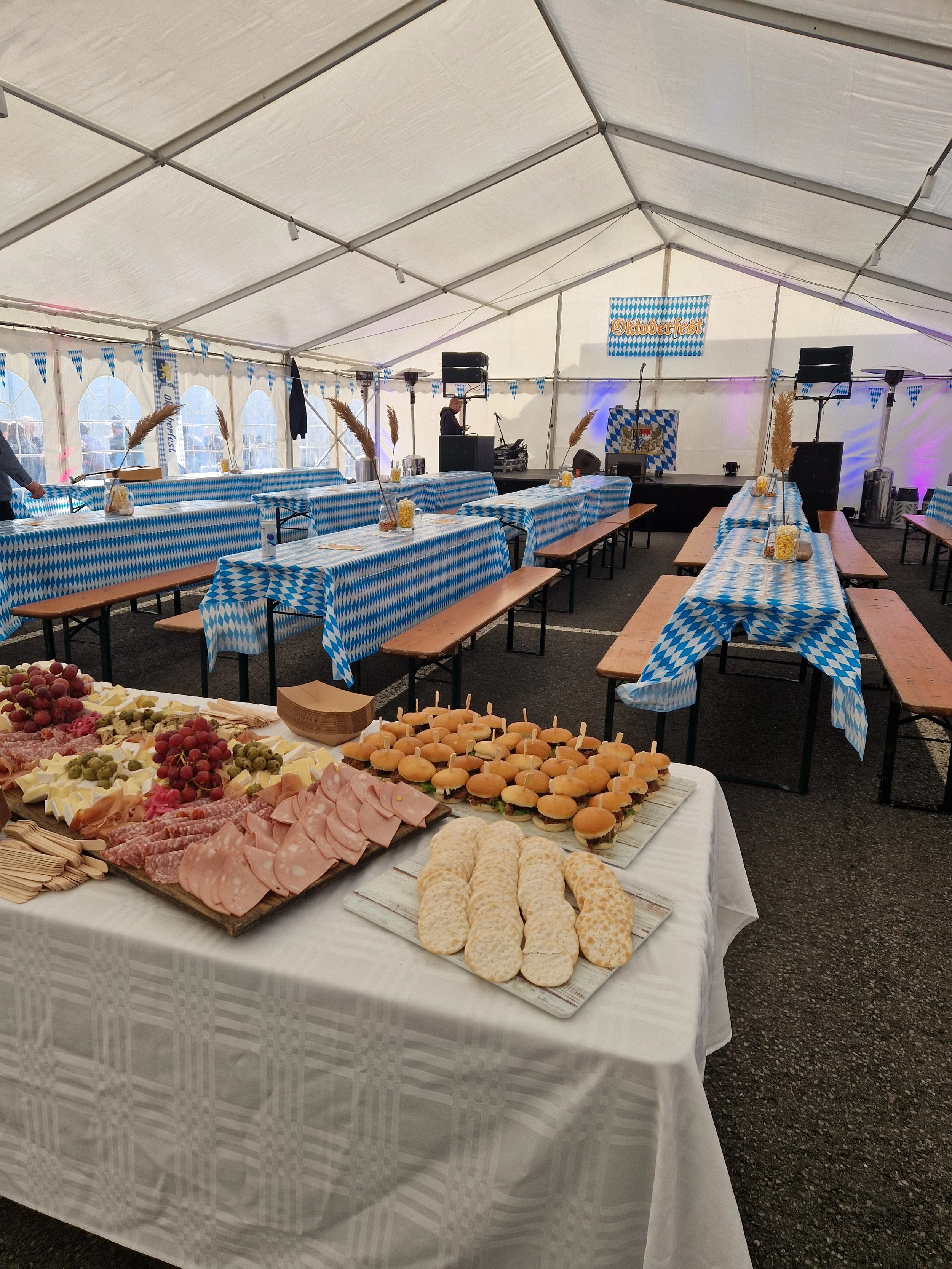 Buffet table with assorted cheeses, meats, grapes, crackers, and small sandwiches, set in a decorated tent for Oktoberfest celebration.