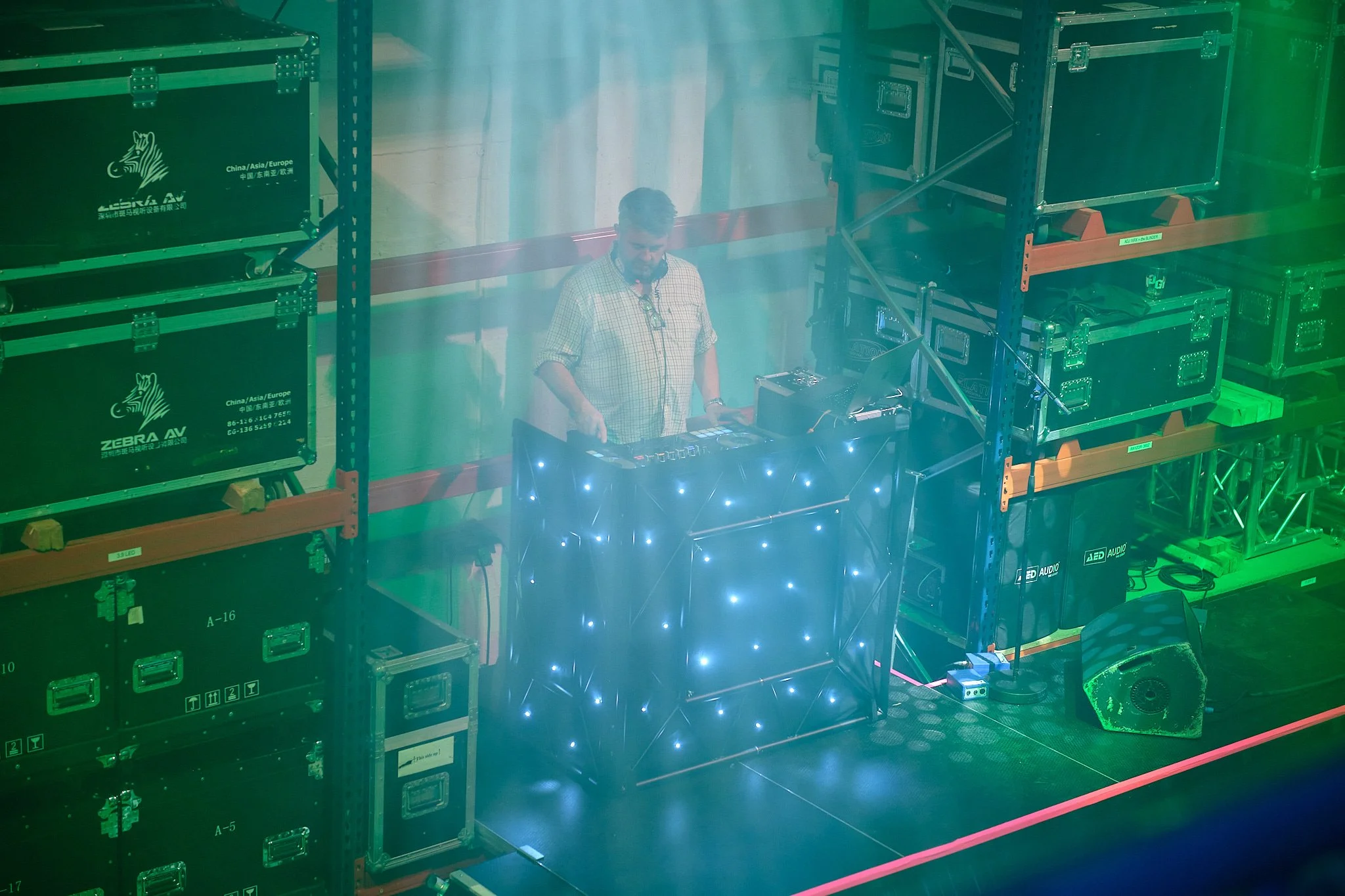 A man operating DJ equipment in a warehouse with black storage boxes on orange shelves, green and blue lighting.