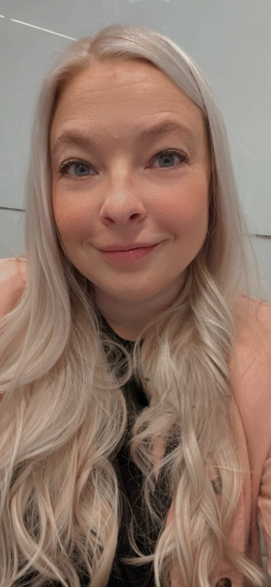 Close-up of a woman with long, wavy platinum blonde hair, smiling softly, wearing a peach-colored jacket, against a plain background.