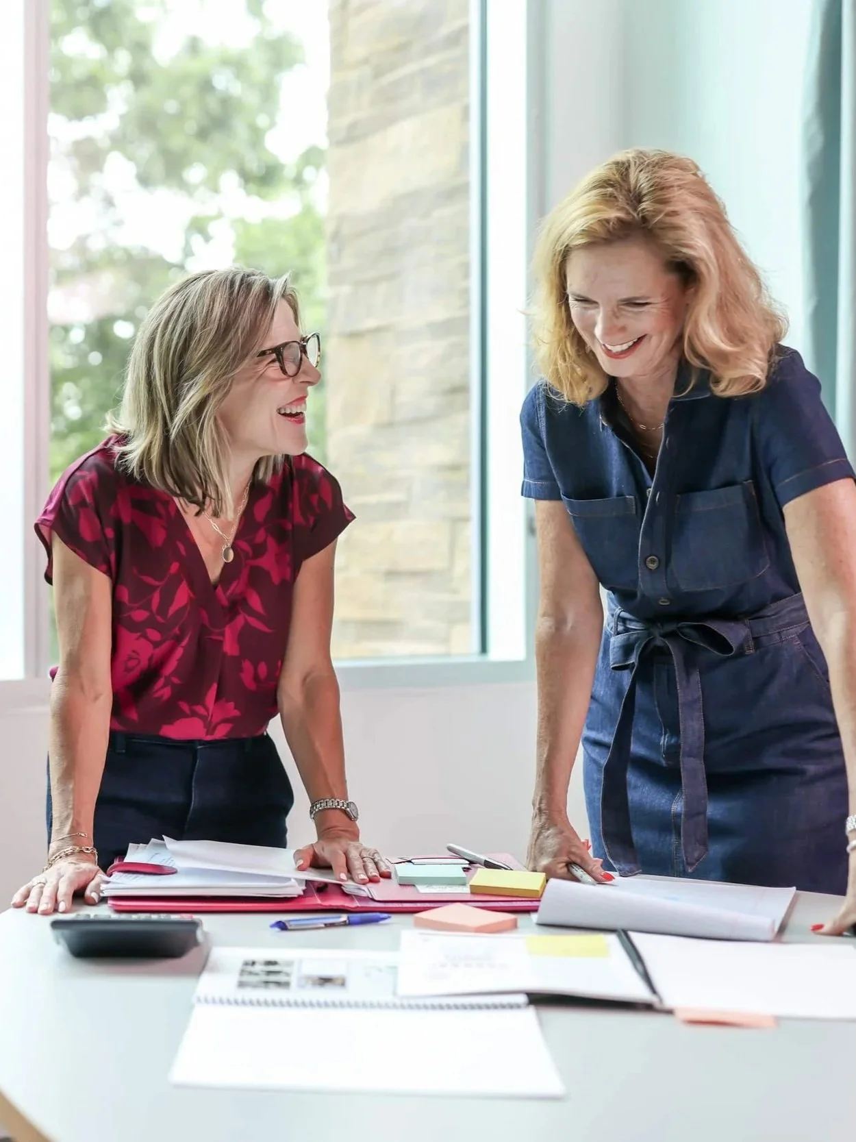 Two women smiling and leaning over a table covered with papers, notebooks, and office supplies, in an office with large windows.