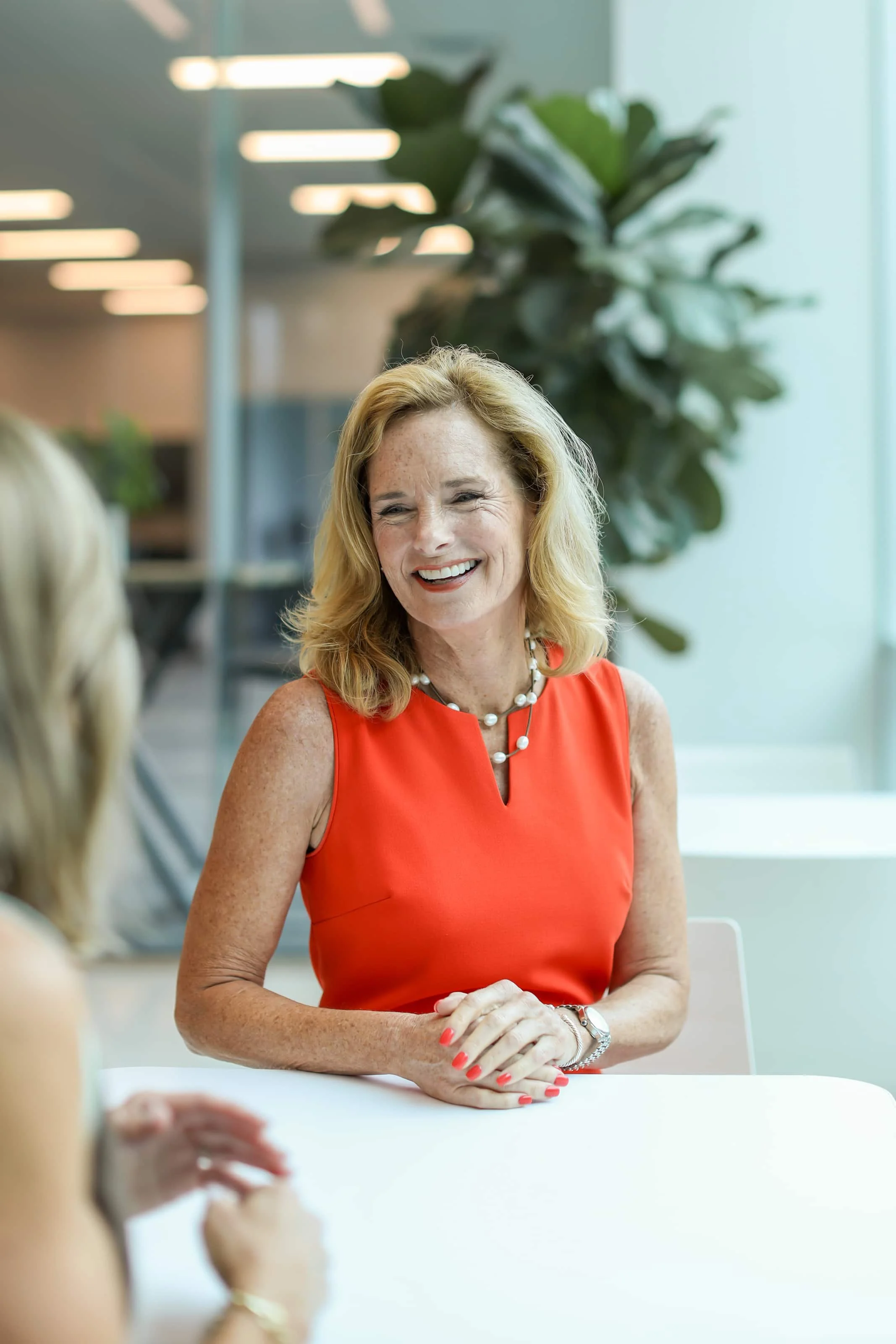 A middle-aged woman with blonde hair, wearing a sleeveless red dress and pearl necklace, smiling and sitting at a table in an office setting.