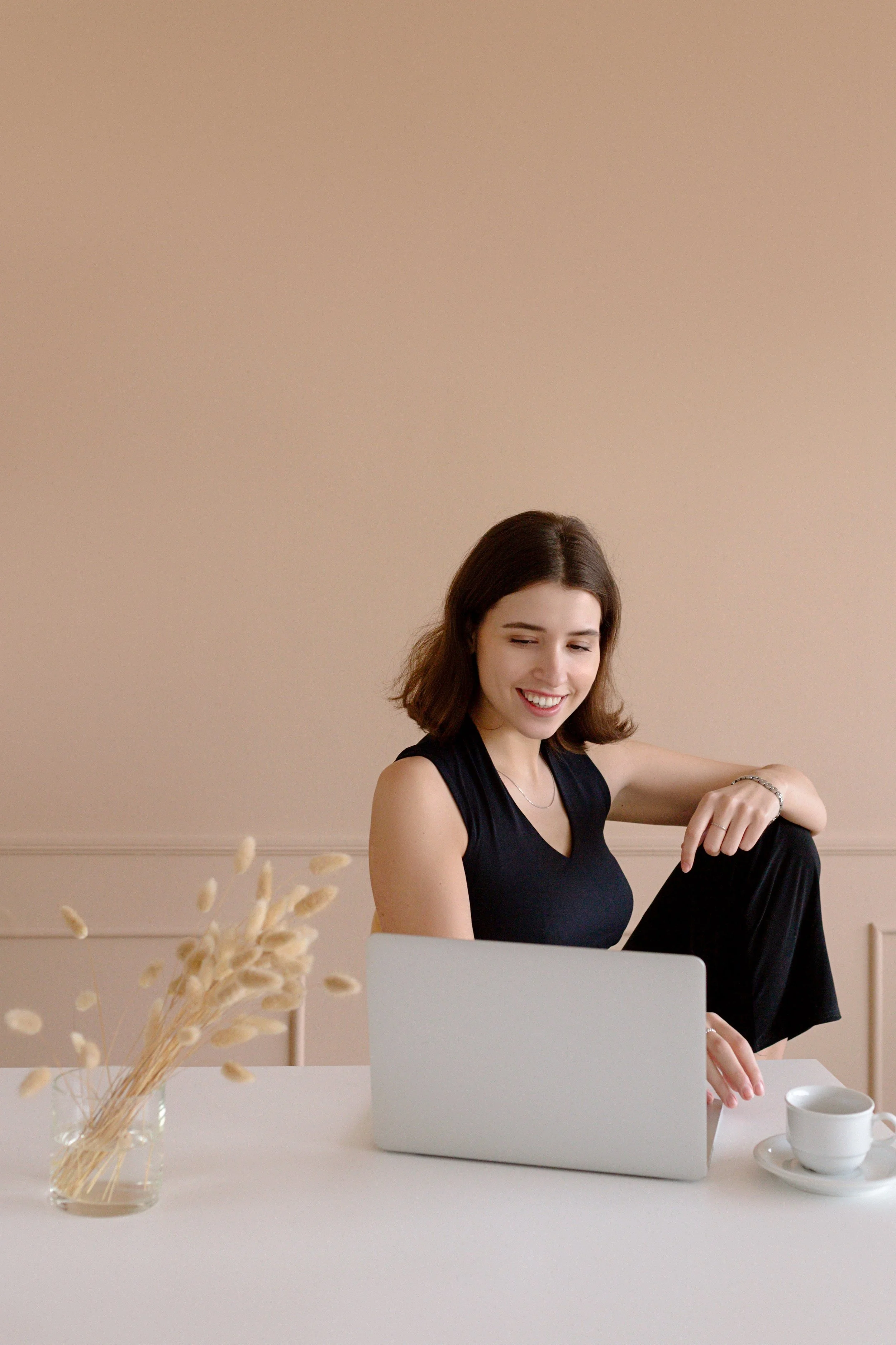 A woman with brown hair sitting at a white table in front of a laptop, smiling and looking down. The background is beige. There is a white cup and saucer on the table, and a glass jar with dried plants to the left of the laptop.