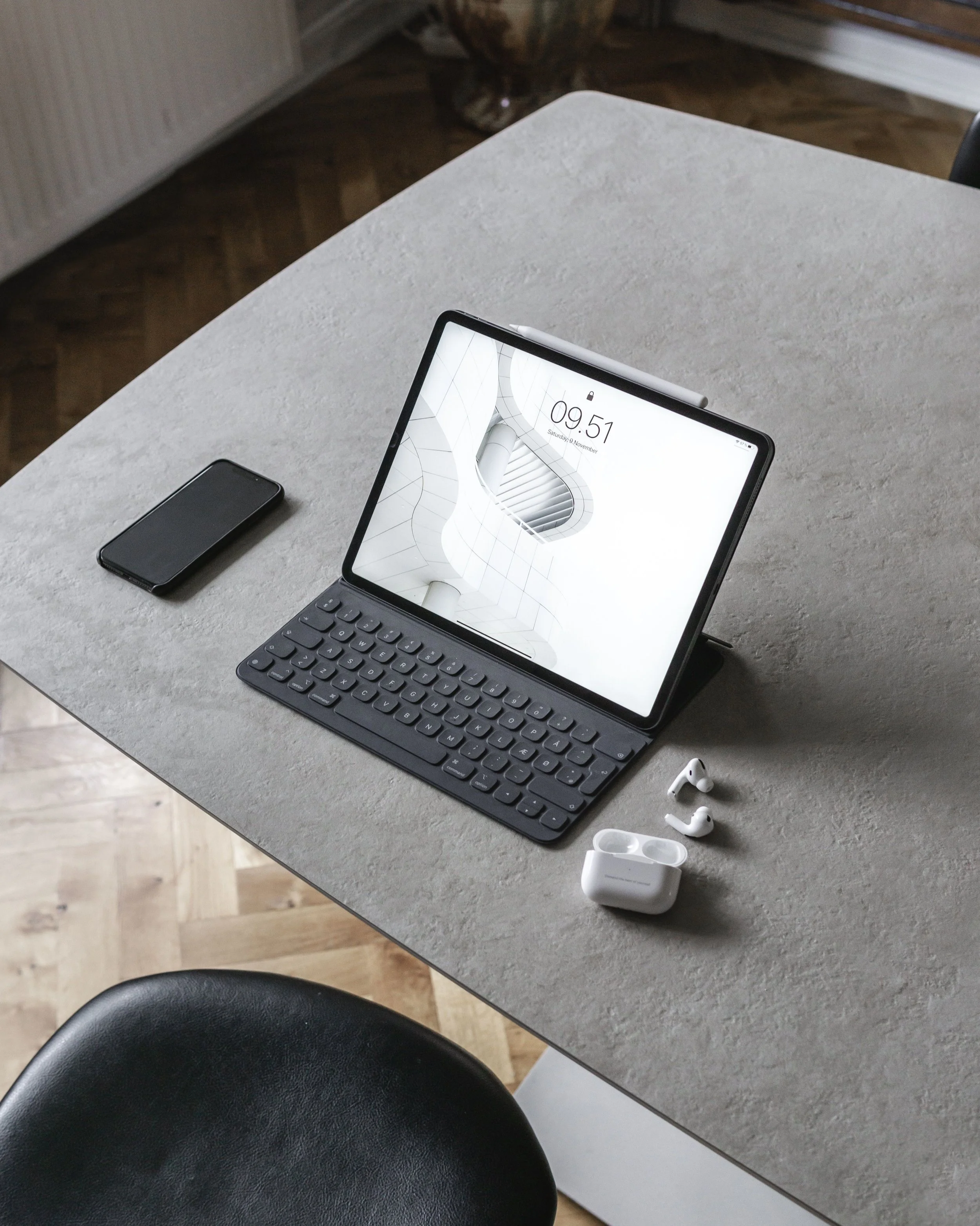 A desk with a tablet on a keyboard stand displaying a modern indoor architectural design, a smartphone to its left, and wireless earbuds with a charging case in front, all on a gray surface in a room with wooden flooring.
