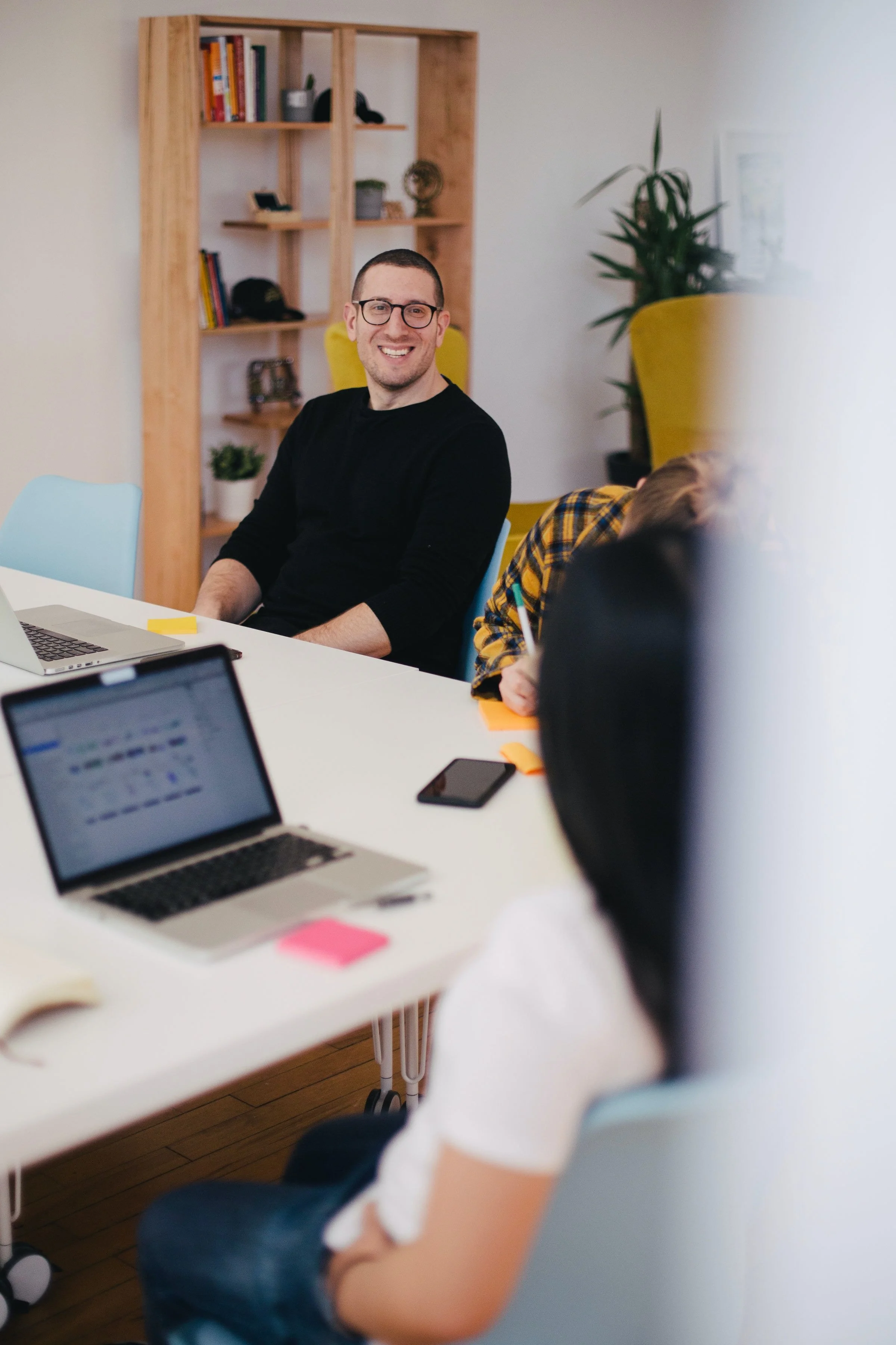 A man with glasses and a black shirt smiling at a meeting table with laptops and notebooks, in a room with wooden floors, a bookshelf, and a yellow chair.
