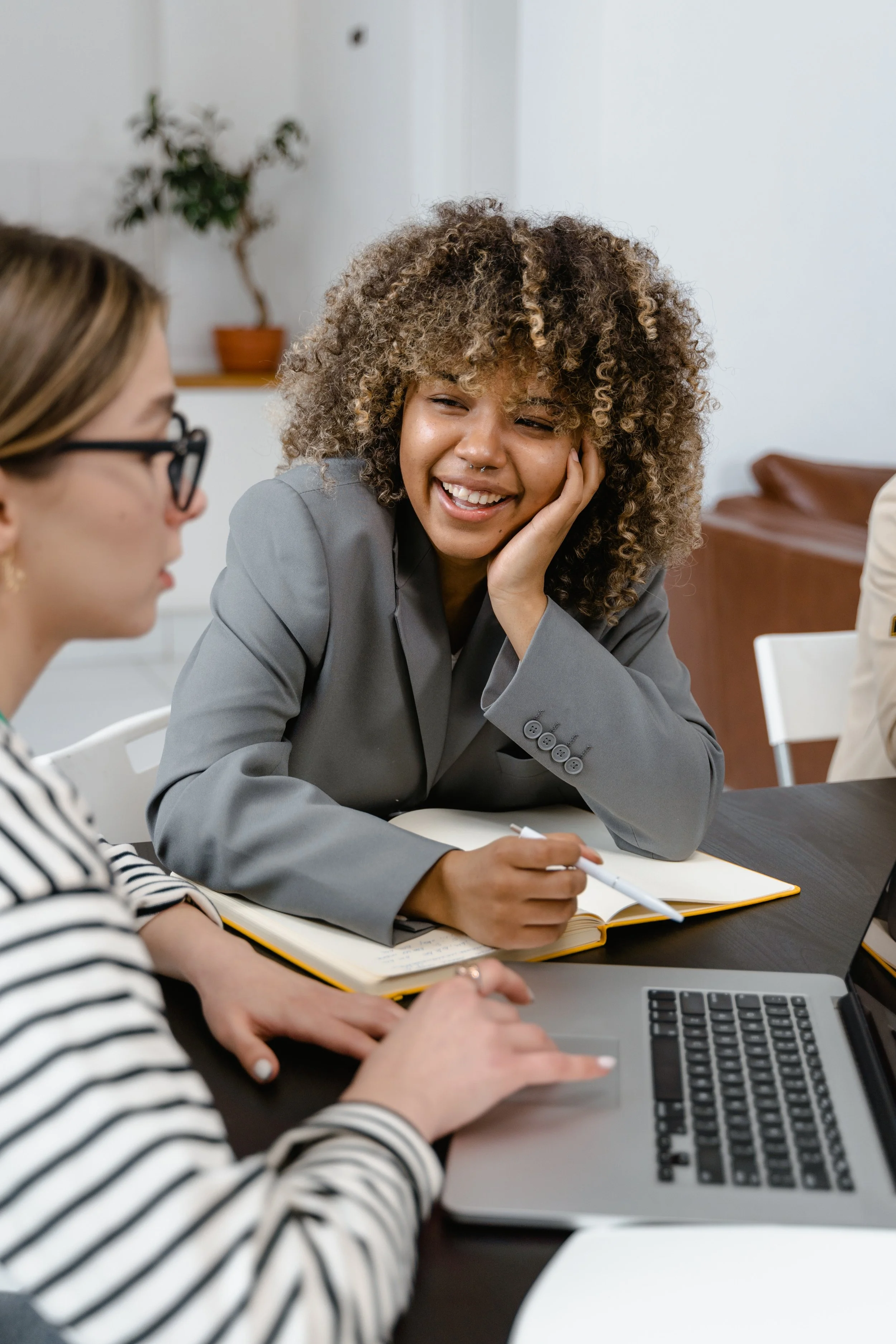 Two women at a meeting, one with curly hair smiling and resting her head on her hand, the other with glasses and striped shirt taking notes with a pen, in a room with a potted plant in the background.