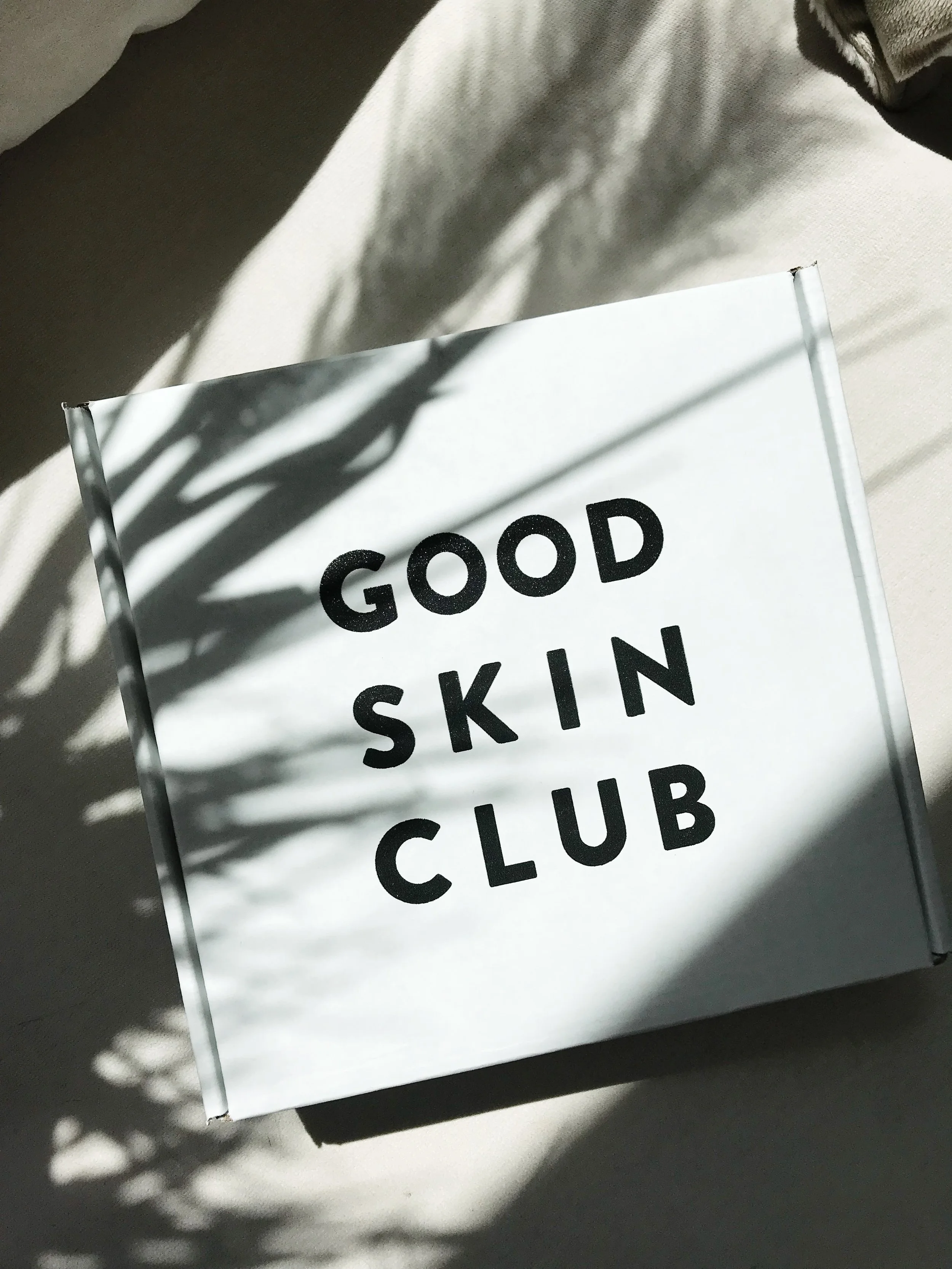 Close-up of a white card with black text reading 'GOOD SKIN CLUB' placed on a light-colored surface with shadows of plant leaves cast over it.
