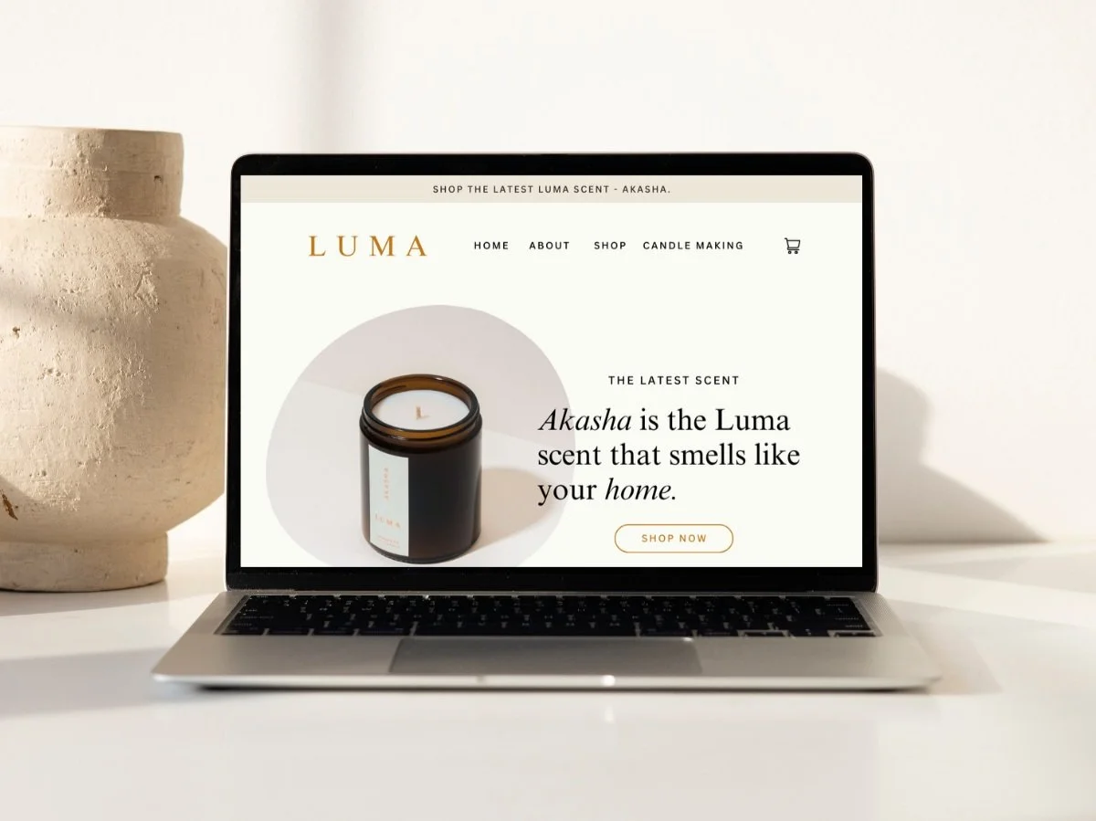 Laptop on a white table displaying a website for Luma scented candles, featuring a candle jar with a black label, text about the Akasha scent, and a "Shop Now" button, with two beige vases on either side.