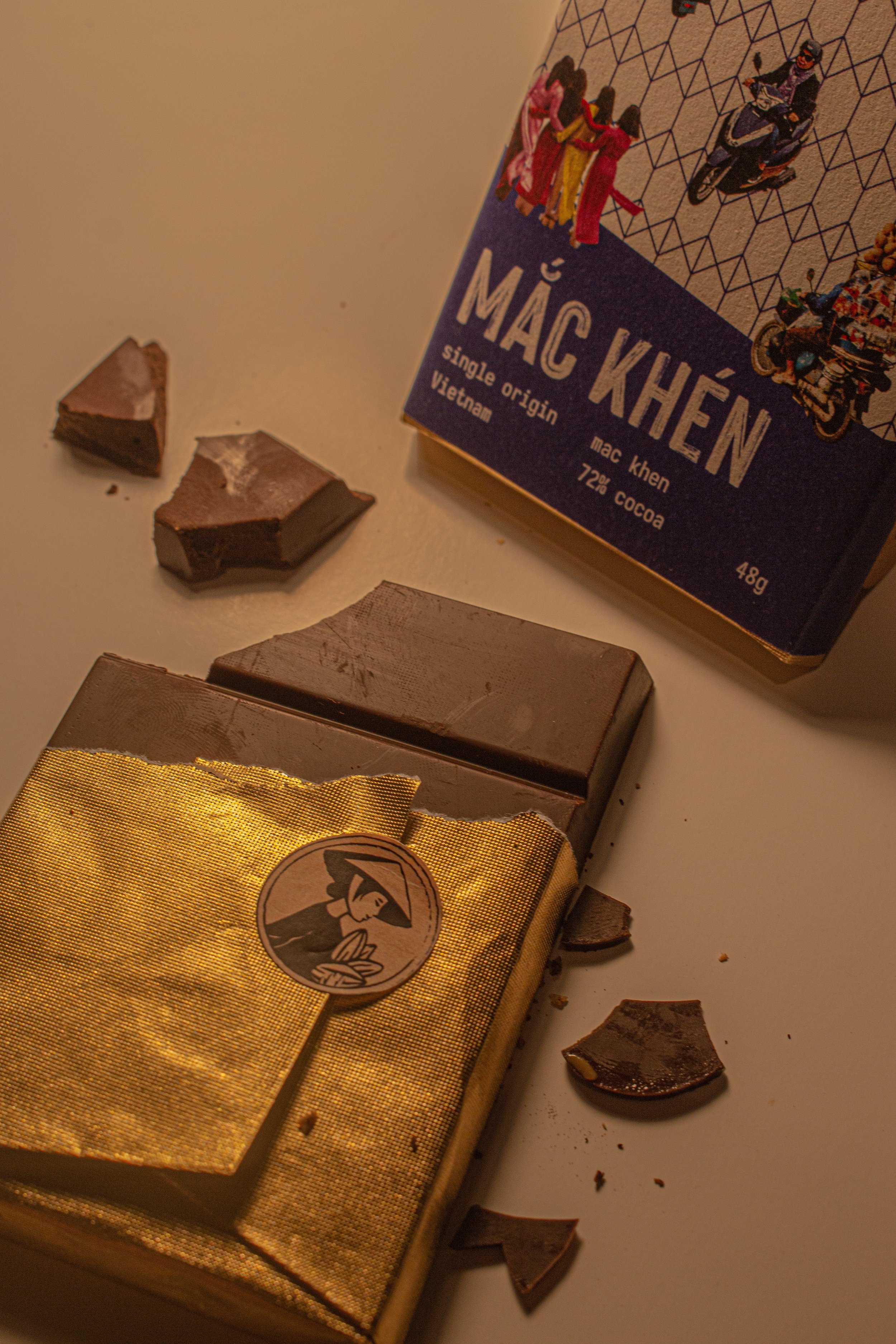 A partially broken bar of chocolate with a gold wrapper, a gold package with a logo, and a box of Mac Khen chocolate bars with Vietnamese writing, depicting scenes of people riding motorbikes and dancing.