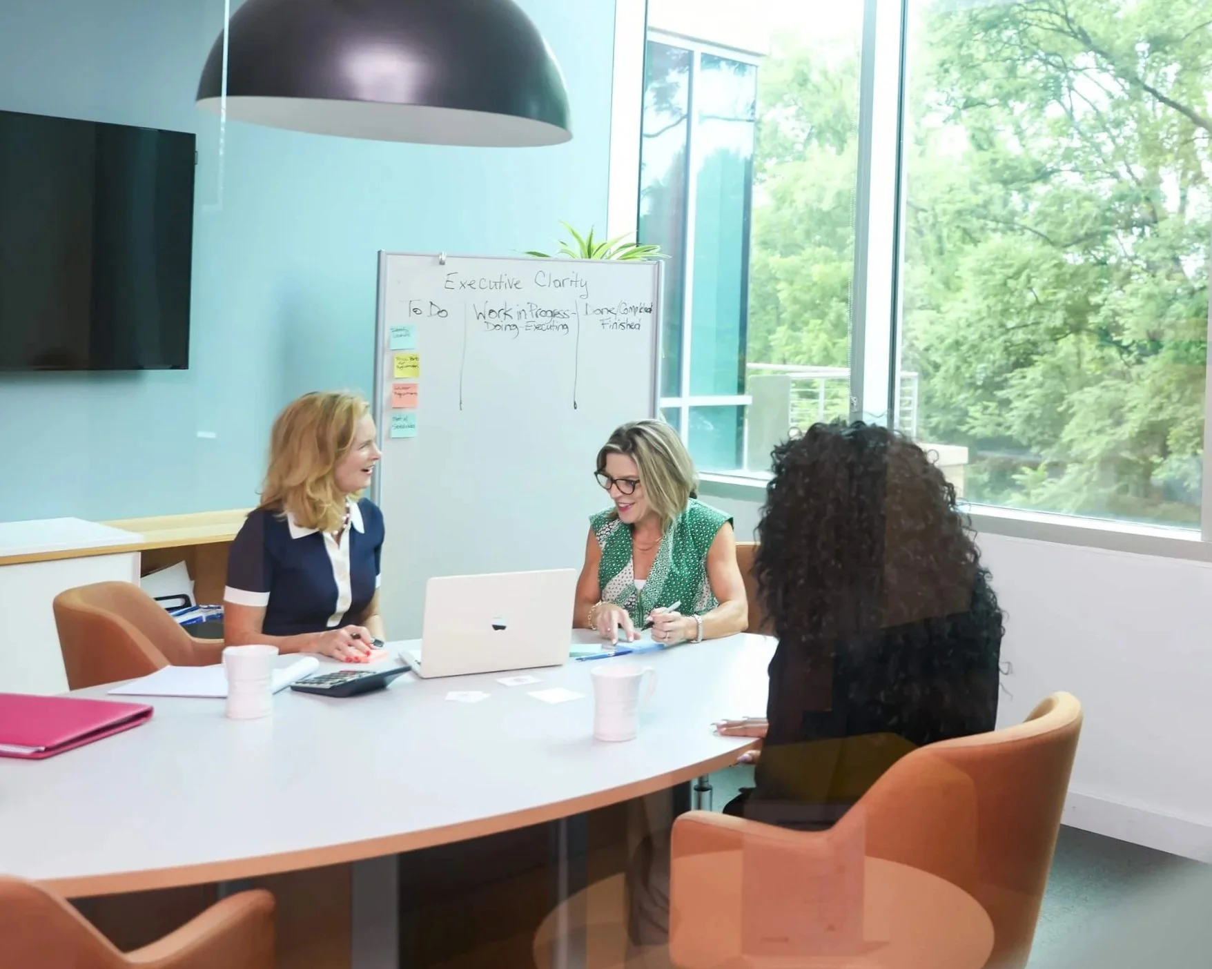 Four women in a meeting room with large windows, a whiteboard, and a wall-mounted TV, engaged in discussion around a table with laptops and papers.
